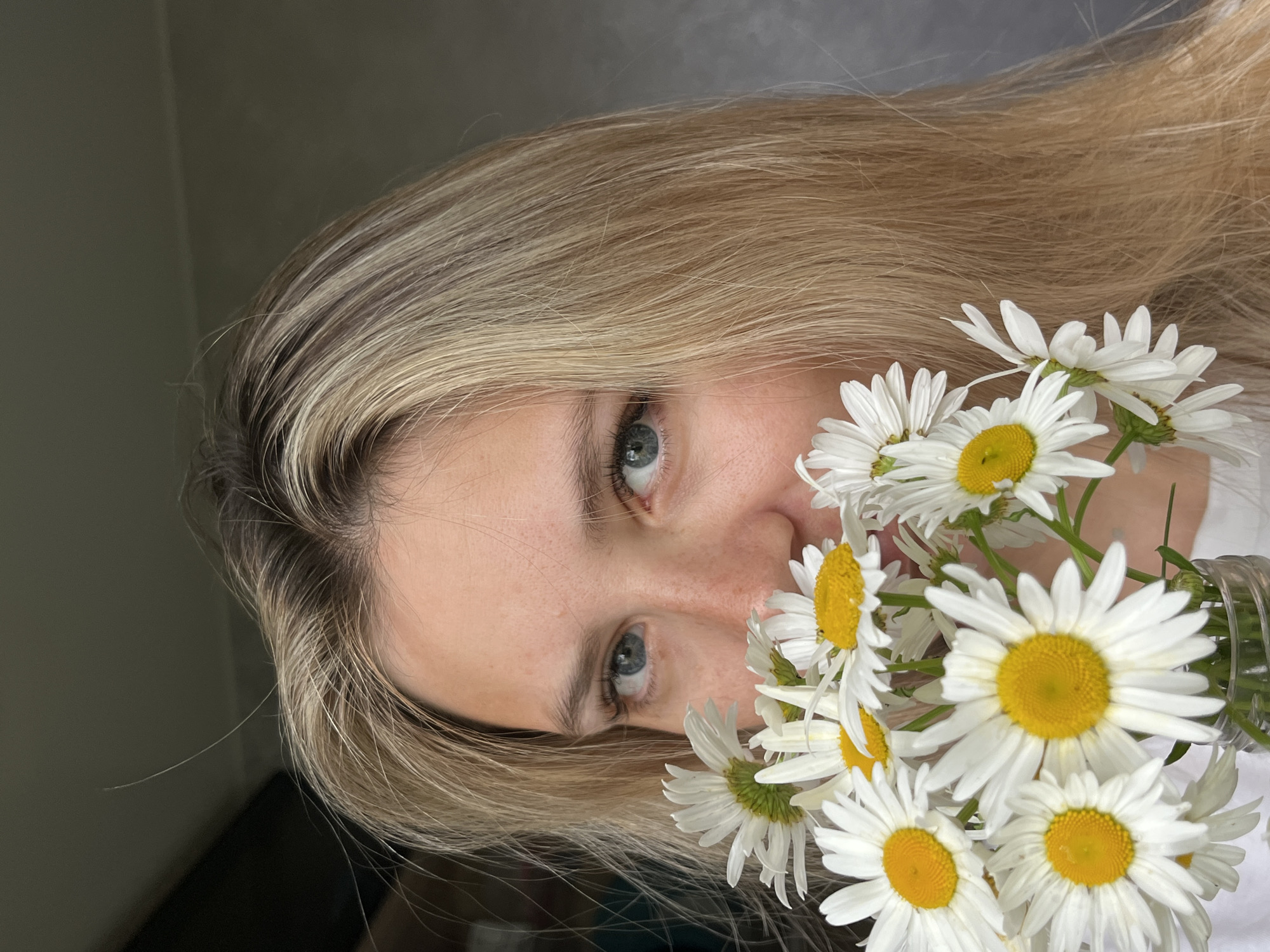 A close-up shot shows a young woman with blonde hair and blue eyes holding a bouquet of white daisies. The flowers partially obscure her face. She is wearing a white shirt. The background is out of focus and appears to be an indoor setting with a neutral-colored wall. There are no visible landmarks or contextual elements that indicate the specific location within Chaikovsky, Russia.