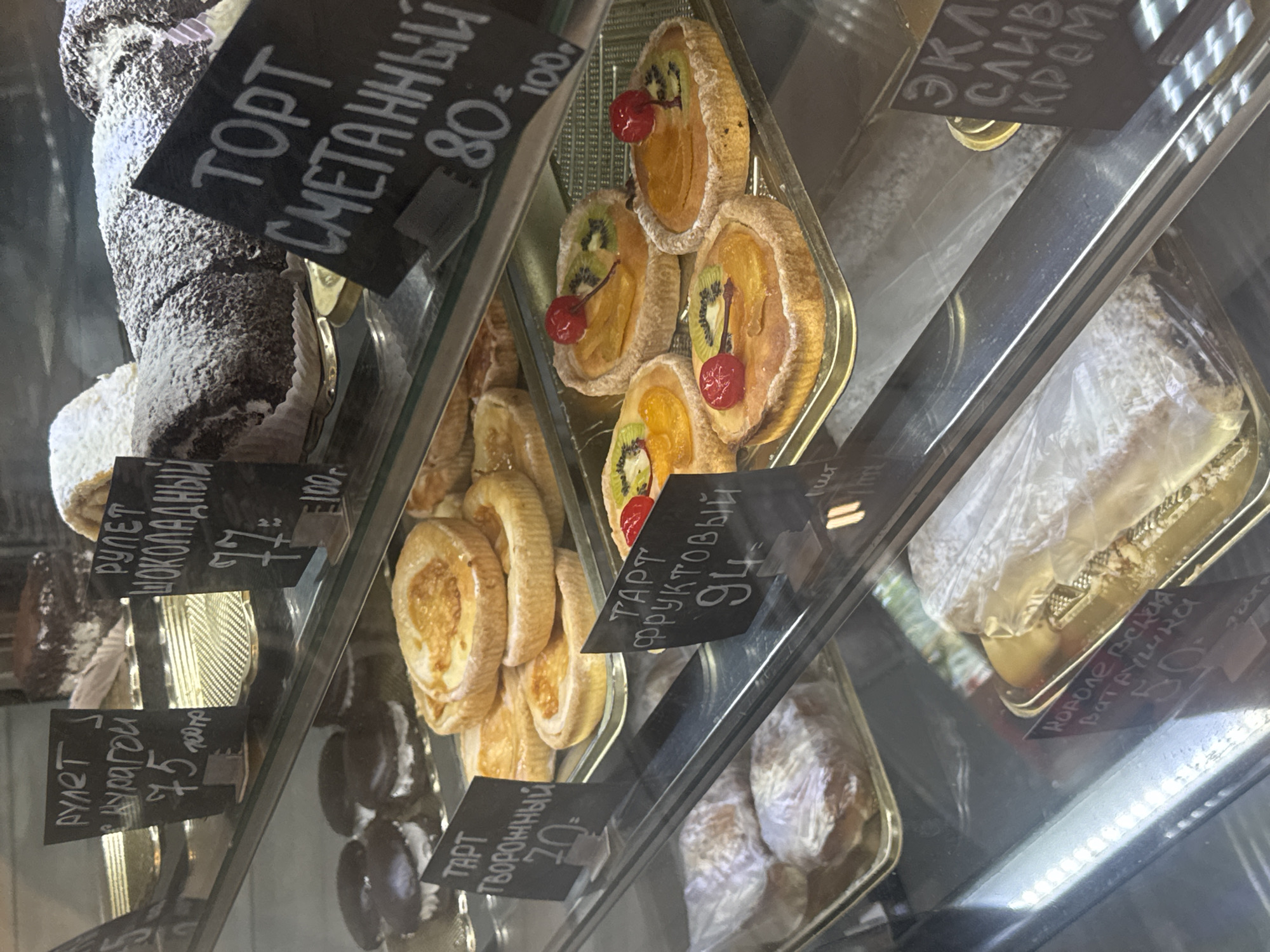 The image shows a display case filled with various pastries in what appears to be a shop or bakery in Chaikovsky, Russia. The pastries are labeled with prices written in Cyrillic script, along with a possible abbreviation or denomination. In the upper left, a tall chocolate-covered cake is labeled "TORT SMETANNYI" and priced at "80-100". Next to it, are fruit tarts with kiwi and cherries. Underneath, there's a chocolate roll labeled "RULET SHOKOLADNYI" for "77". Other pastries include tarts with apricot filling, priced at "75", another tart with a curd filling for "70", and a pastry called "KOROLEVSKAYA VATRUSHKA" priced at "50". The environment is a typical bakery or pastry shop setting, with the focus on displaying a variety of baked goods for sale.
