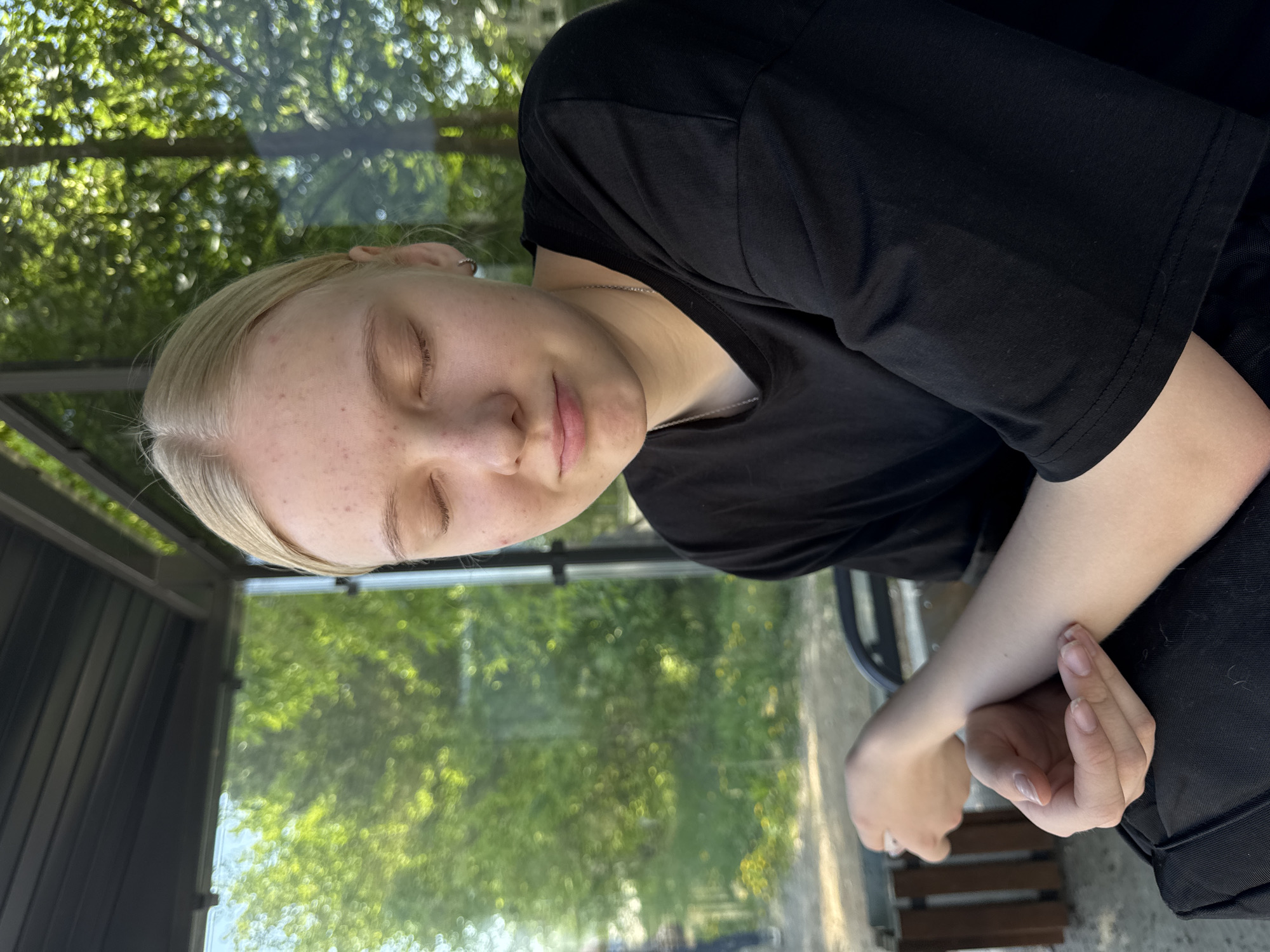 A young, fair-skinned woman with blonde hair is sitting with her eyes closed. She is wearing a black t-shirt. Her hands are folded in her lap. She appears to be sitting in a glass-enclosed bus stop. Behind the glass, there is greenery suggesting trees. The location is Chaikovsky, Russia.