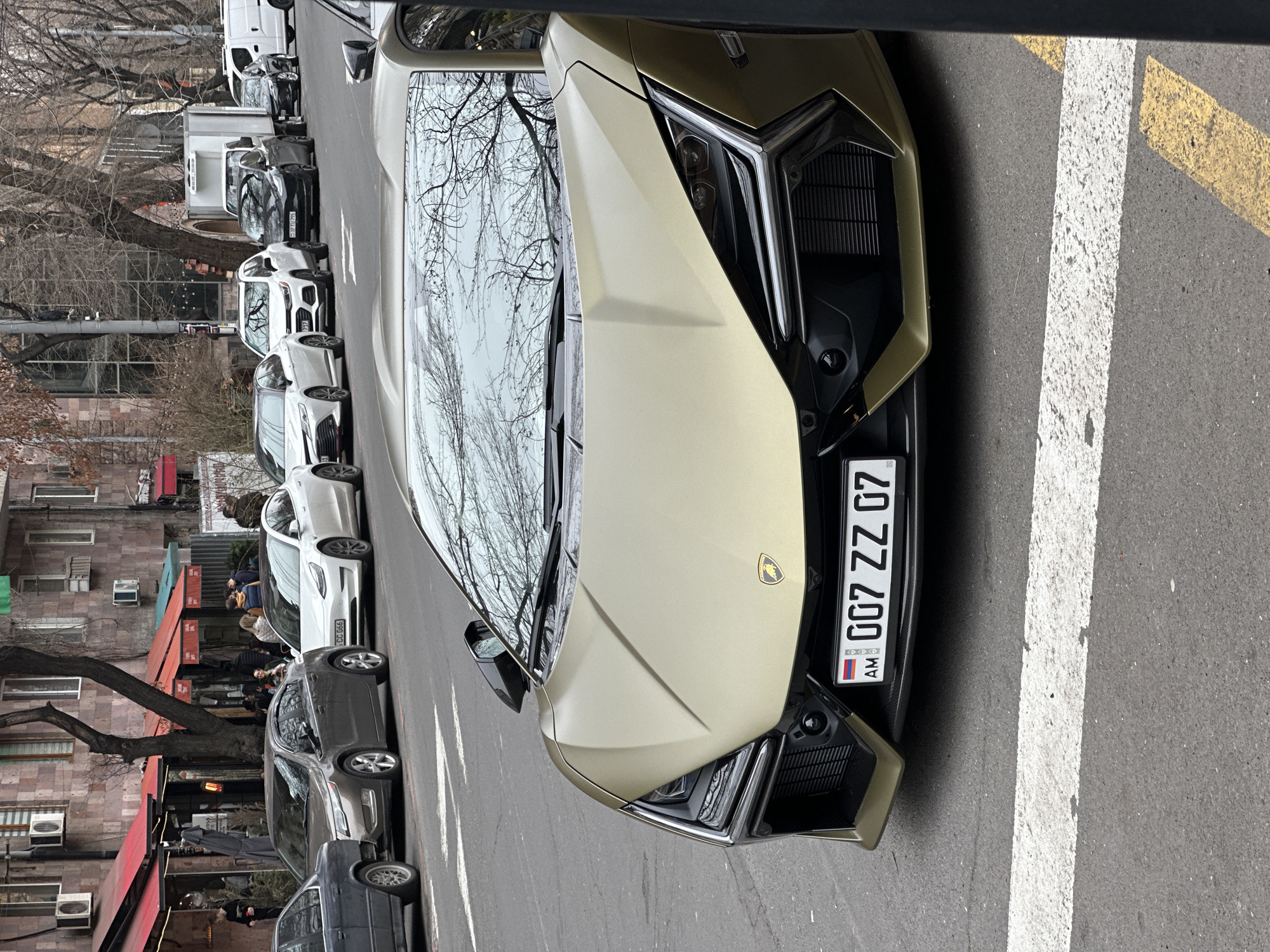 The image displays a street scene in central Yerevan, Armenia, characterized by multi-story buildings constructed from reddish-brown stone, featuring multiple windows and exterior air conditioning units. The foreground is dominated by a gold/bronze matte finish Lamborghini Sian FKP 37, parked facing forward on the asphalt street. The vehicle bears an Armenian license plate, "AM 007 ZZ 07," prominently displayed on its front bumper. White crosswalk lines are visible on the road surface in front of the car.
To the left of the Lamborghini, a line of various vehicles is parked parallel to the curb, including a dark grey sedan, a dark brown Chevrolet Volt, a white Tesla Model 3, a white Toyota Camry, a white Chevrolet Trax, a black Tesla Model 3, a white box truck, and a white cargo van. Beyond these vehicles, several commercial establishments with red awnings are visible at ground level. Multiple individuals are present outside these businesses, some seated at outdoor tables and others standing, engaged in unspecified activities. Bare deciduous trees with intricate branch patterns line the street and are reflected in the vehicle windshields. The overall lighting suggests an overcast daytime scene during a cooler season.