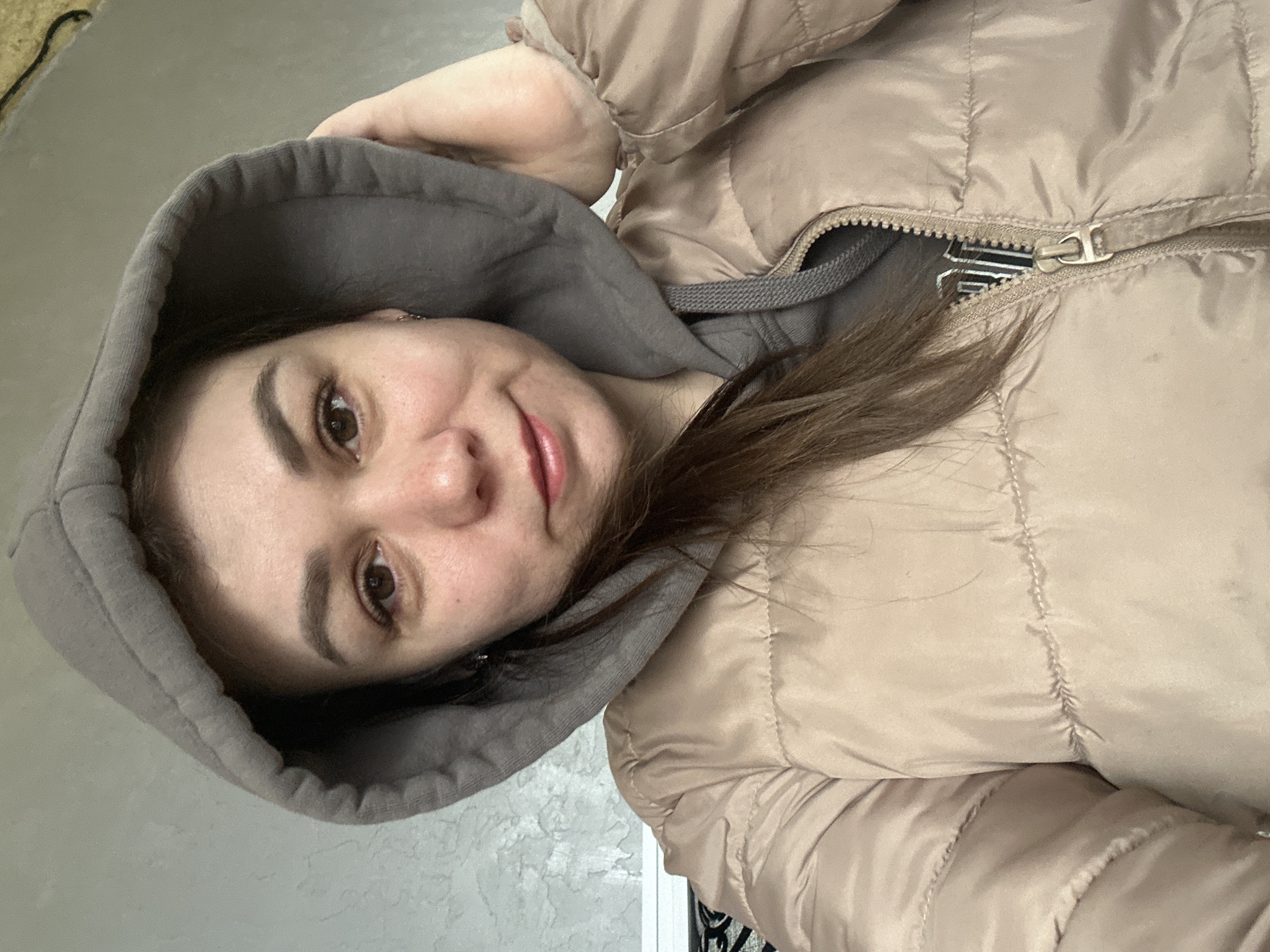 A woman with fair skin and brown hair is wearing a beige quilted jacket with a gray hoodie. The jacket is partially zipped up, revealing a striped garment underneath. The background appears to be a textured, light-colored wall, possibly indoors. The overall impression is a self-portrait or selfie, with the camera angled slightly downward toward the subject. The specific location is not visually discernible from the image; hence, it is impossible to confirm whether it is within the city of Chaikovsky, Russia.
