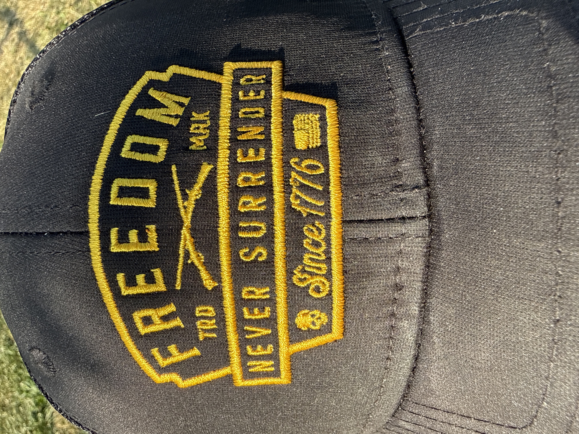 A close-up, high-angle shot shows a black baseball cap with a yellow embroidered patch. The patch features the word "FREEDOM" in large yellow letters across the top, with "TRD" and "MRK" below it. Below that, in a rectangular box, is the phrase "NEVER SURRENDER". At the bottom of the patch, "Since 1776" is written, with a small skull symbol to the left and what appears to be a historical image of two figures to the right. The cap is positioned outdoors, with a blurred background of green and brown grass.