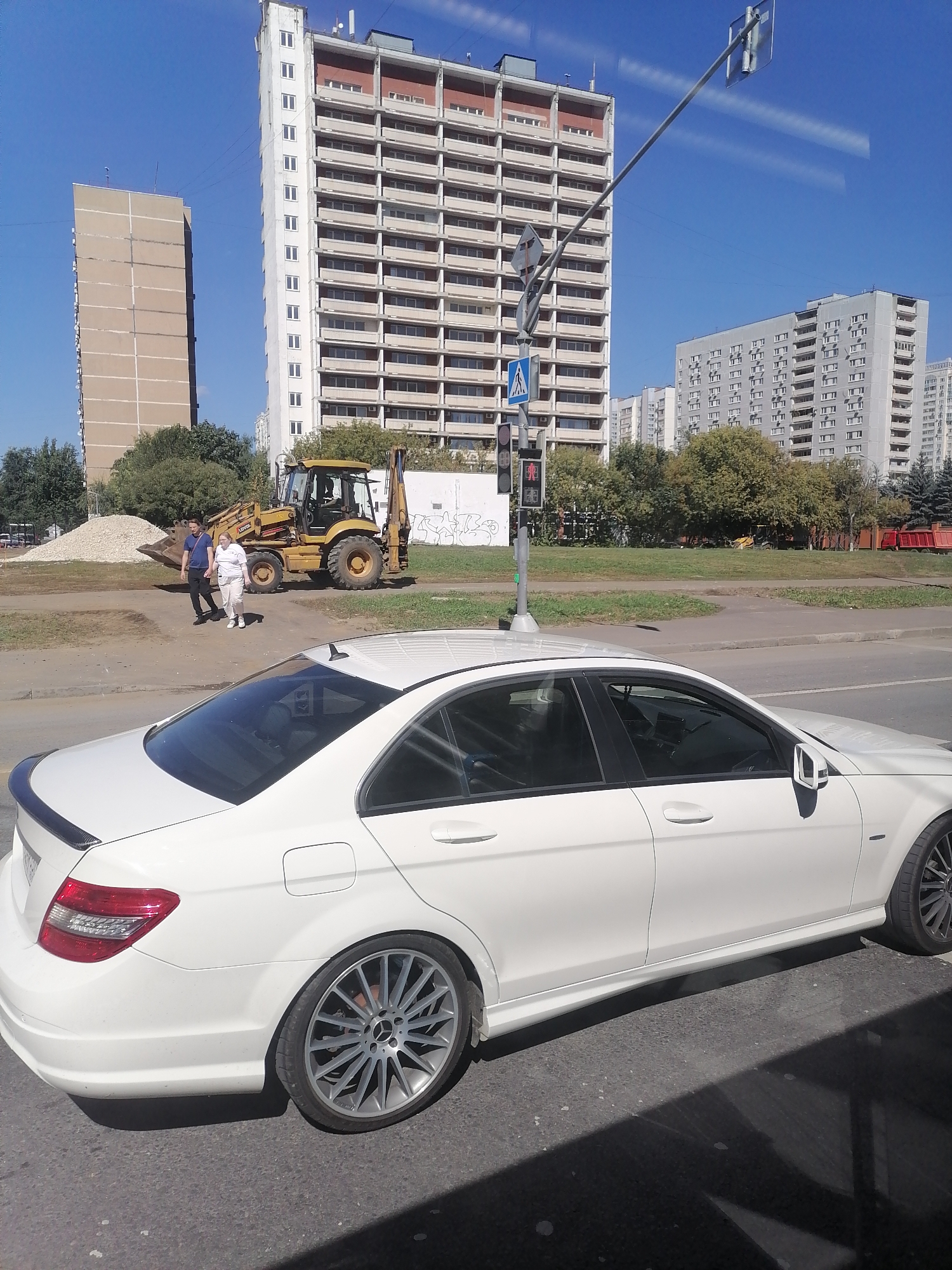 In Moscow, Russia, on a sunny day, a white Mercedes sedan is parked on the right side of the road, partially obscuring the view of the background. In the background, a yellow backhoe loader is parked on a dirt area. Two people, a man and a woman, are walking from the backhoe loader towards the left side of the image. Behind them, a large pile of gravel is visible. Further in the background, several apartment buildings and a traffic signal with a pedestrian crossing sign are present. The sky is clear and blue.