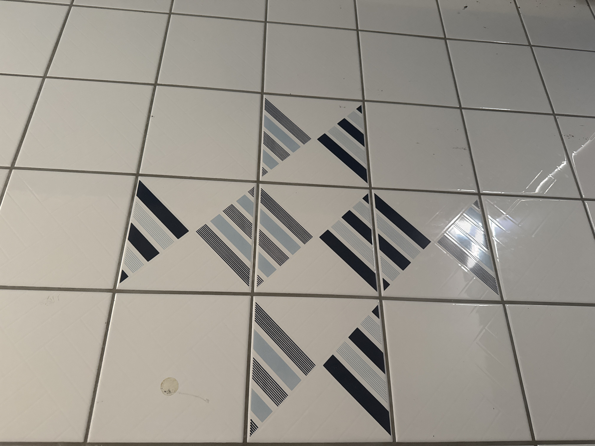 The image shows a section of a tiled wall in Le Landeron, Switzerland. The tiles are square and white with gray grout lines. Some of the tiles have a decorative pattern consisting of diagonal stripes in navy blue and light blue. There are also some small specks and smudges on the tiles, suggesting they are not new. The lighting in the scene appears to be artificial, possibly from overhead lights, as evidenced by reflections on the glossy tile surfaces. The overall impression is of an indoor wall, perhaps in a bathroom or kitchen.