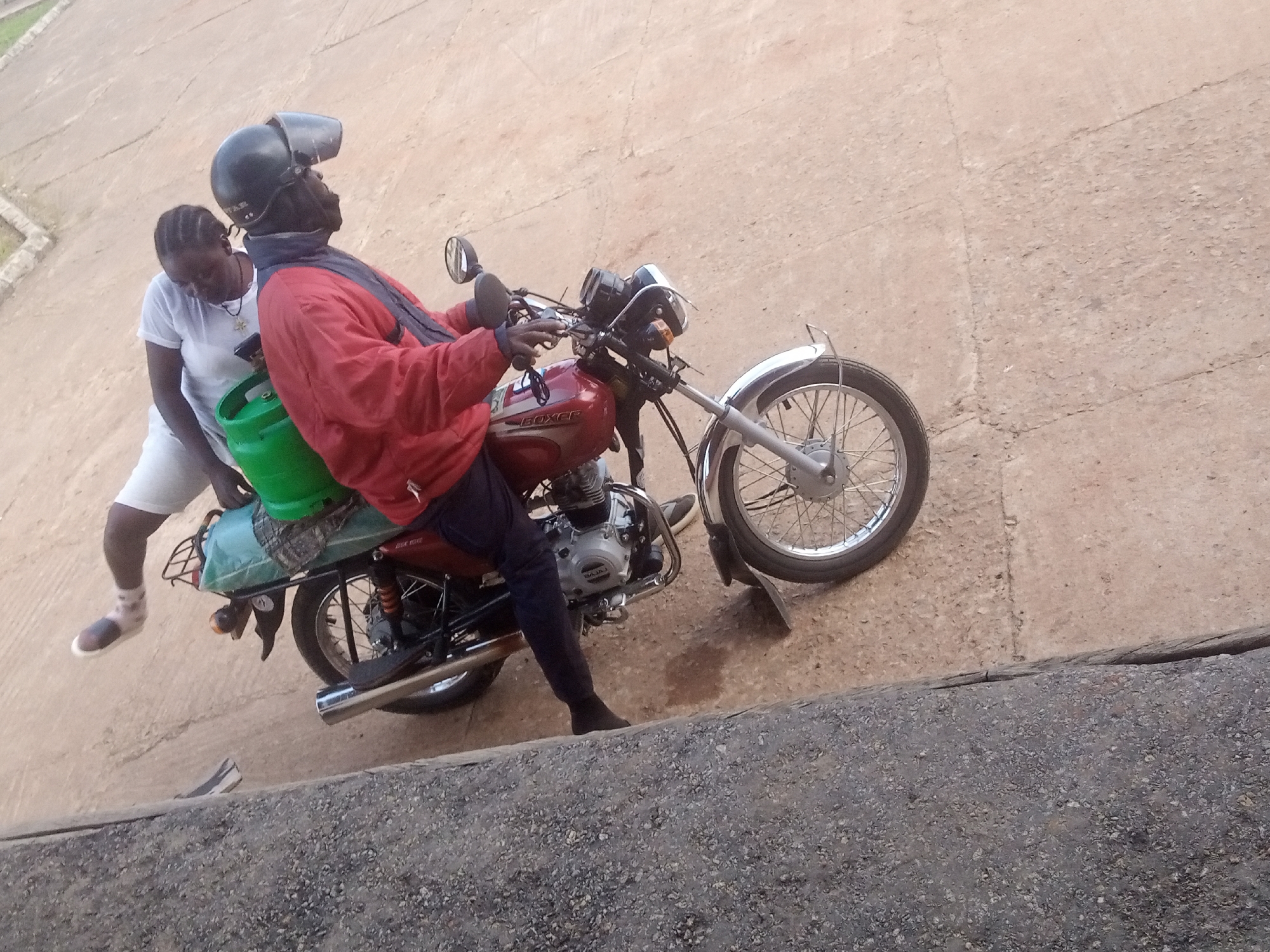 Two people, a man and a woman, are on a motorcycle. The man is driving, wearing a red jacket, dark pants, and a black helmet. The woman is a passenger behind him, wearing a white shirt and white shorts. A green water jug is strapped to the back of the motorcycle. The motorcycle is dark red and silver, with chrome accents. The scene appears to be outdoors, on a paved surface that looks like a road or a courtyard. The ground is a light brown, cracked concrete. The lighting suggests it's daytime, possibly late afternoon due to the angle of the light. There is no visible text in the image.