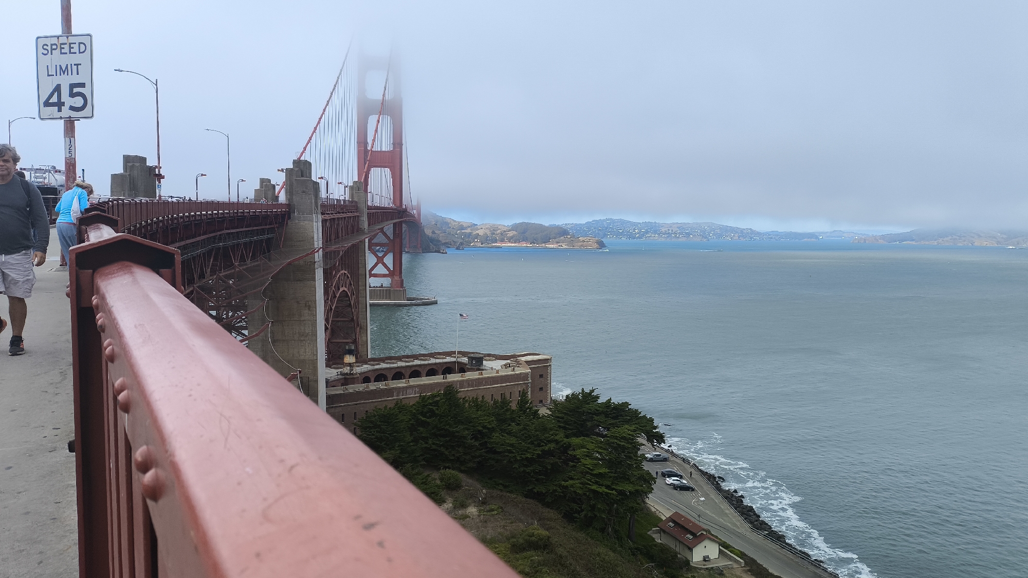 The image shows a view from the Golden Gate Bridge in San Francisco, California. On the left, a man in a dark shirt and light shorts walks on the bridge's sidewalk. To his right, a person in a blue top and light pants stands looking out. The iconic red suspension bridge is visible, with its towers disappearing into the fog. Below the bridge, a historical fort structure with arched openings and trees lining its edge is situated on the coastline. A road runs alongside the water, with parked cars. The vast expanse of the bay stretches towards the horizon, with distant hills and islands shrouded in mist. The sky is overcast.