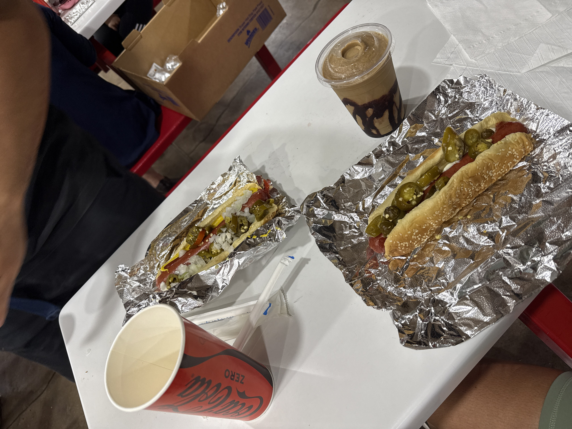 The image shows a white table at what appears to be a food court or casual dining area. On the table, there are two hot dogs wrapped in aluminum foil, a cup of Coca-Cola Zero, a plastic cup containing a blended drink (likely coffee or chocolate), and some wrapped straws. The hot dogs are loaded with toppings including jalapeños, onions, and mustard. The table is next to a red chair. Two people are seated at the table, with one wearing a black shirt and dark shorts, and the other wearing a green shirt. Based on the products and style of food, it is likely that this scene is at a Costco food court. The location within Puebla, Mexico cannot be determined from the images provided.
