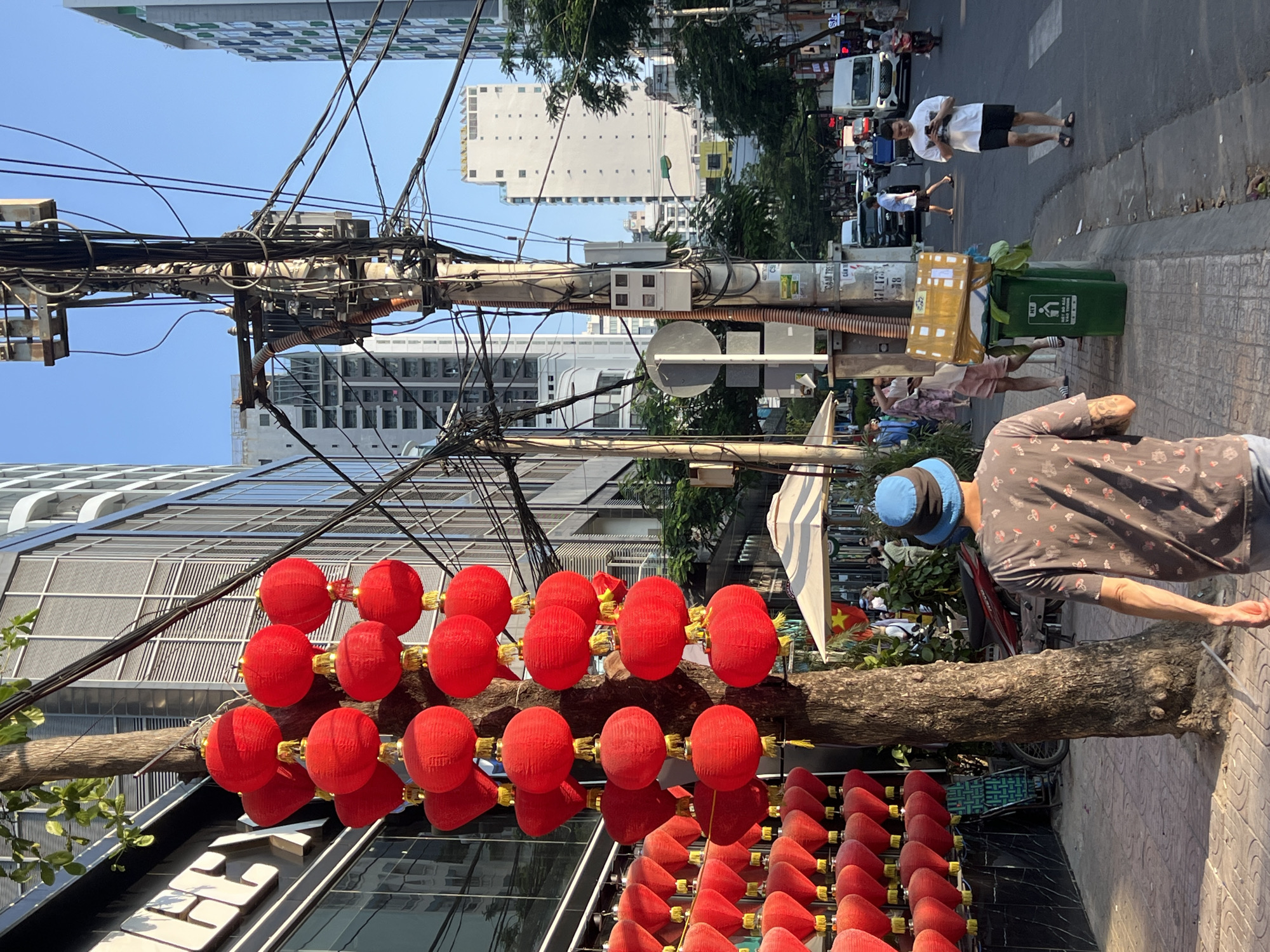 The image shows a street scene in Nha Trang, Vietnam. Red lanterns are strung across a tree branch that is attached to the front of a building. Overhead, a utility pole is cluttered with wires. A man wearing a blue bucket hat and a patterned shirt stands on the sidewalk next to the tree. Behind him, a green trash can overflows with a cardboard box and plants. In the background, pedestrians walk on the sidewalk and across the street amid traffic. Buildings of varying heights line the street, with the sky visible in the upper portion of the frame.