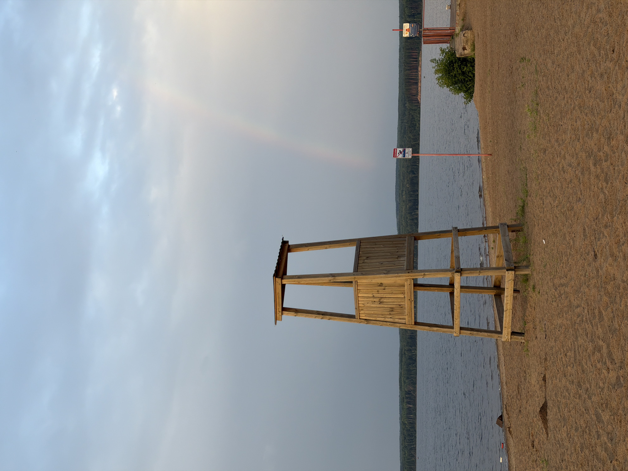The image shows a sandy beach next to a body of water, likely a river or lake, with a dense forest lining the opposite shore. In the foreground, a wooden lifeguard tower stands prominently on the sand. To the right of the tower, two red posts with signs are visible near the water's edge. One sign appears to be a warning sign with a pictogram. A faint rainbow is visible in the cloudy sky above the water, suggesting recent or ongoing rain. The overall scene is serene, with no people or activity depicted.
This scene is located at the Kama River embankment in Chaikovsky, Russia, a popular recreational area known for its beaches and forest-lined shores. The lifeguard tower is a common fixture at such locations, ensuring safety for swimmers. The presence of warning signs indicates designated areas or rules for water activities. The natural beauty of the landscape, with the water, forest, and the ephemeral appearance of a rainbow, creates a picturesque setting.