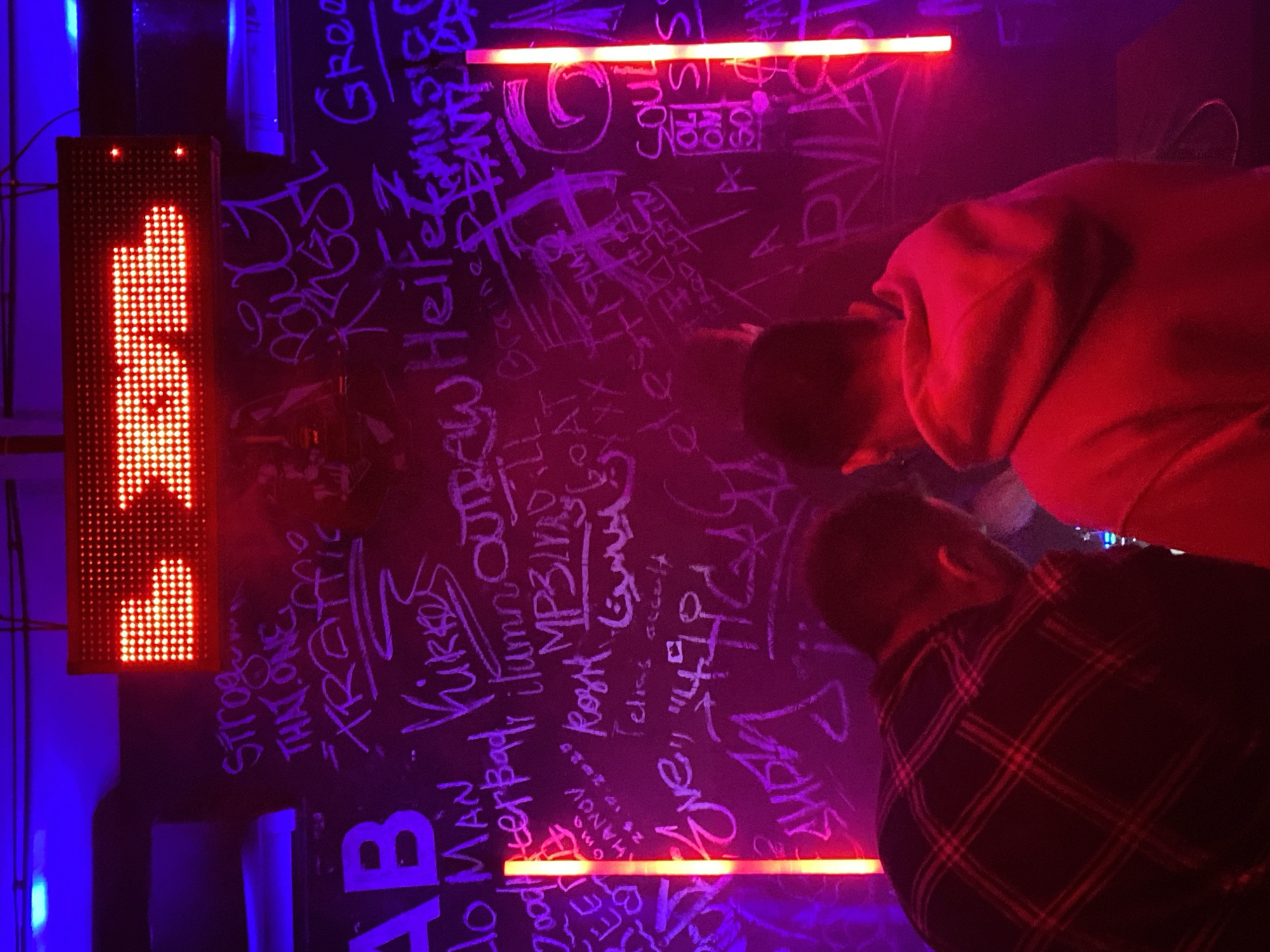Two individuals are standing in front of a wall covered in white graffiti under red and blue lighting. One person is wearing a red hooded sweatshirt, while the other is in a dark plaid shirt. Next to the graffiti wall is a vertical red LED sign displaying an unidentifiable word. The location appears to be in an urban setting, likely a bar or club in Unknown, Unknown, as suggested by the environment and lighting.