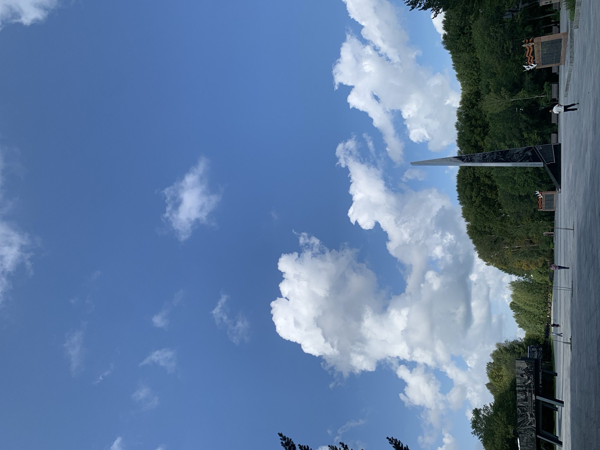 The image depicts the Victory Park in Prokopyevsk, Russia, on a day with a bright blue sky dotted with fluffy white clouds. In the foreground, a paved open area is visible with a few people walking. To the right, a tall, abstract monument with a metallic finish stands prominently, adorned with what appear to be historical etchings or reliefs. To the left, another exhibit or structure with a dark, angular design and etched imagery is visible. Behind these monuments, a dense line of green trees forms a backdrop, with some lighter-colored foliage interspersed, suggesting a mix of tree species or seasonal changes. The overall scene is one of an outdoor memorial park.