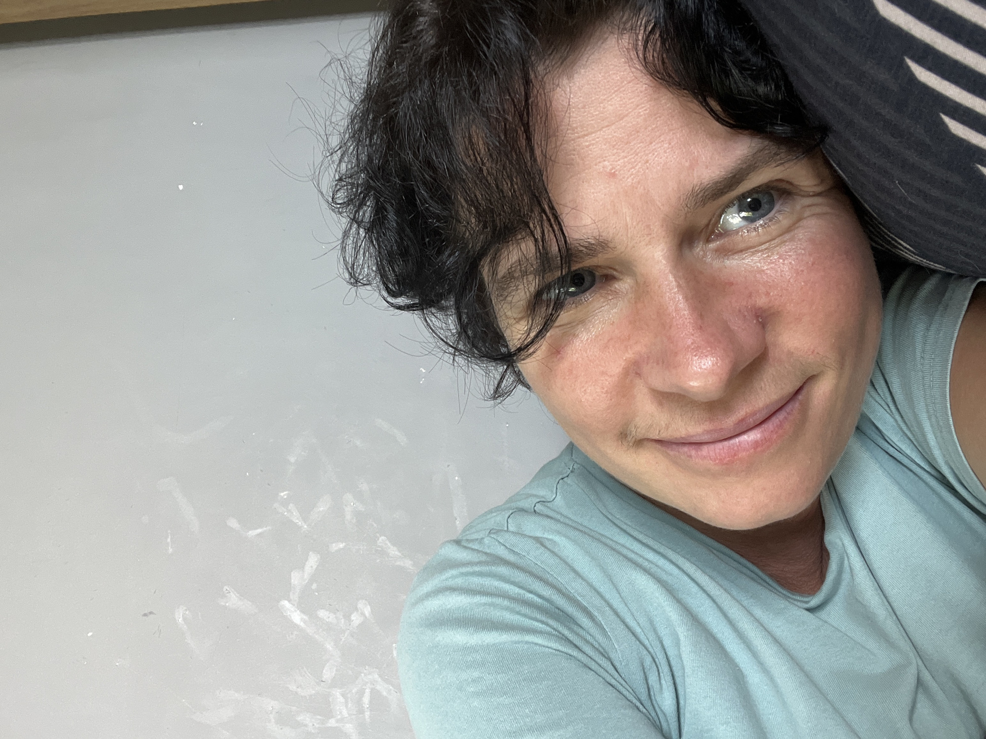 A close-up, overhead shot shows a person resting on a pillow, their head tilted and their body angled to the left. They are wearing a light blue t-shirt. Their face, which is partially illuminated, shows visible skin texture, including some redness around the cheeks and eyes. A slight smile is visible on their lips. Their dark, curly hair is scattered around their head, and one blue eye is clearly visible. The background consists of a light-colored wall with some white, smudged marks. The scene is likely indoors, possibly in a bedroom or similar private space, given the presence of a pillow and the relaxed posture of the individual. There is no identifiable information within the image or metadata to confirm the location as Ureki, Georgia.