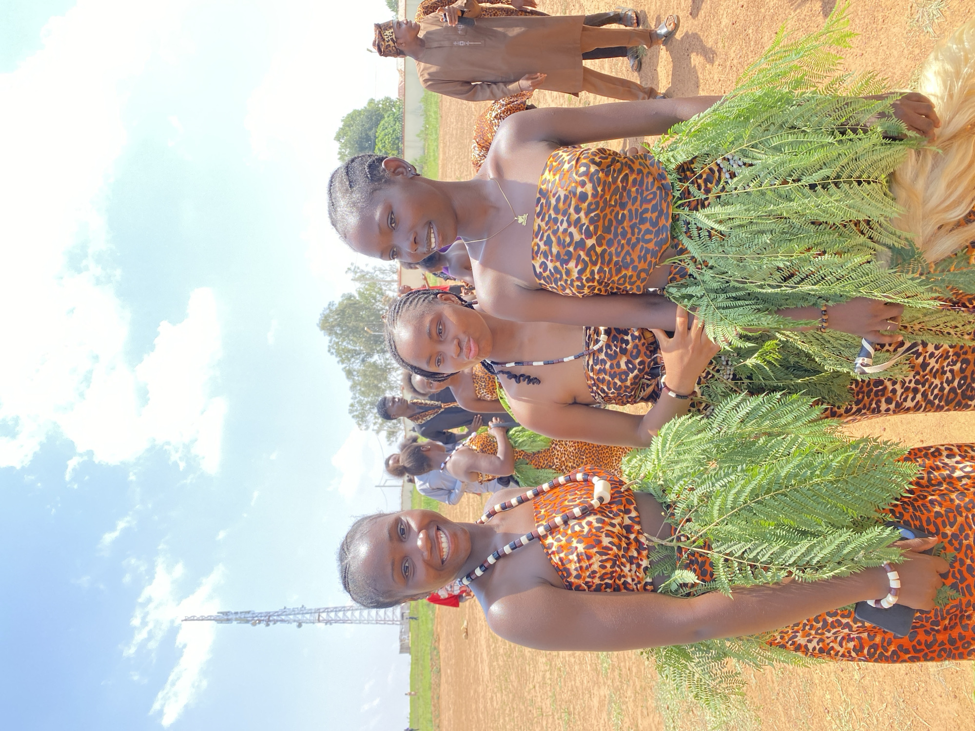 Three young women are prominently featured in the foreground of the image, each wearing a leopard-print garment and holding green branches. They are smiling and appear to be posing for the camera. The women are also adorned with necklaces, and have braided hair. Behind them, several other people are visible, some similarly dressed in leopard print, and others wearing casual attire. The setting appears to be an outdoor event, possibly a cultural celebration, taking place on a field of reddish-brown dirt. In the background, there are trees, a building, and some type of metal scaffolding or tower structure. The sky is visible with a mix of blue and white clouds. The specific location is Durimi, Nigeria.