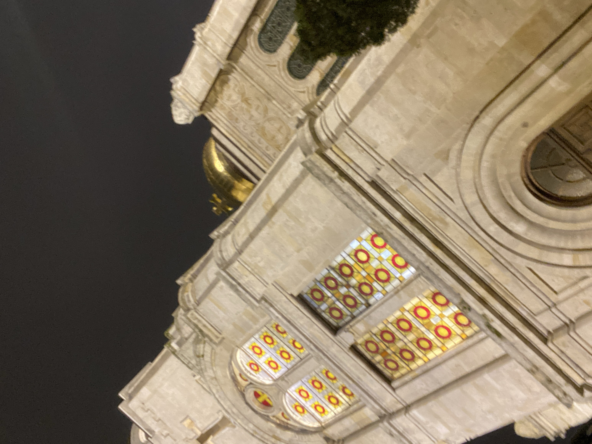 A low-angle, upward shot depicts the exterior of a building at night, with the sky appearing dark. The building is constructed of light-colored stone and features multiple windows with stained-glass panes of geometric patterns with red and yellow circular designs. A golden dome is visible above the building. The architecture includes arched details and ornate carvings. A patch of greenery is seen on top of the building. The building is the Dormition of the Mother of God Cathedral located in Varna, Bulgaria.