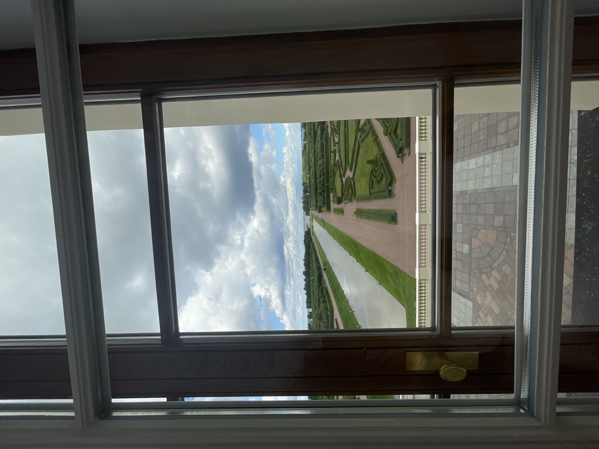 This image captures a view through a window overlooking a vast formal garden and canal in Strelna, Russia. The window frame, made of dark wood with brass hardware, bisects the scene. Outside, a long, reflective canal stretches into the distance, flanked by meticulously manicured green hedges forming geometric patterns in the garden. Beyond the garden, trees line the horizon under a cloudy sky. The foreground visible through the window shows a paved courtyard with square stone tiles. The overall impression is one of grandeur and historical significance, characteristic of imperial estates.