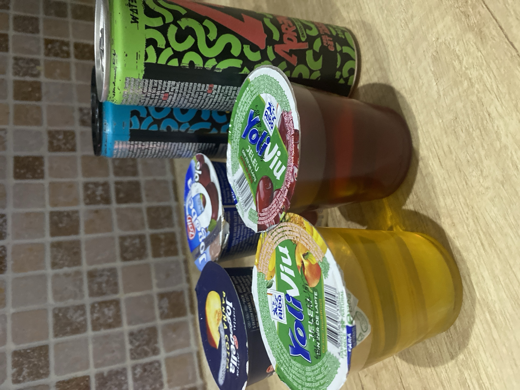 The image depicts a collection of food and beverage items arranged on a wooden surface, likely a countertop or table. In the foreground, two clear plastic cups contain desserts. The cup on the left holds a yellow, gelatinous dessert with a peach-colored fruit slice on its lid. The cup on the right contains a dark red, gelatinous dessert topped with cherries.
Behind the desserts, several containers are visible. To the left, there are two small yogurt cups with blue lids. One yogurt cup has a lid featuring a peach slice. To the right of the yogurts, two beverage cans stand upright. The can on the left is predominantly blue with black graphics. The can on the right is a vibrant green with black and red graphics, bearing the text "WATER".
The background consists of a tiled wall with square tiles in varying shades of brown and beige. The overall lighting suggests an indoor setting. The scene is devoid of any people or overt actions. The objects appear to be stationary, implying a static composition for the photograph.
While the image itself does not contain specific indicators of its location in Chișinău, Moldova, the presence of products like "YoliViu" jelly and "Jogobella" yogurt, which are popular brands in Romania and Moldova, suggests a plausible origin in the region. The Romanian language text visible on the packaging further supports this geographical context.