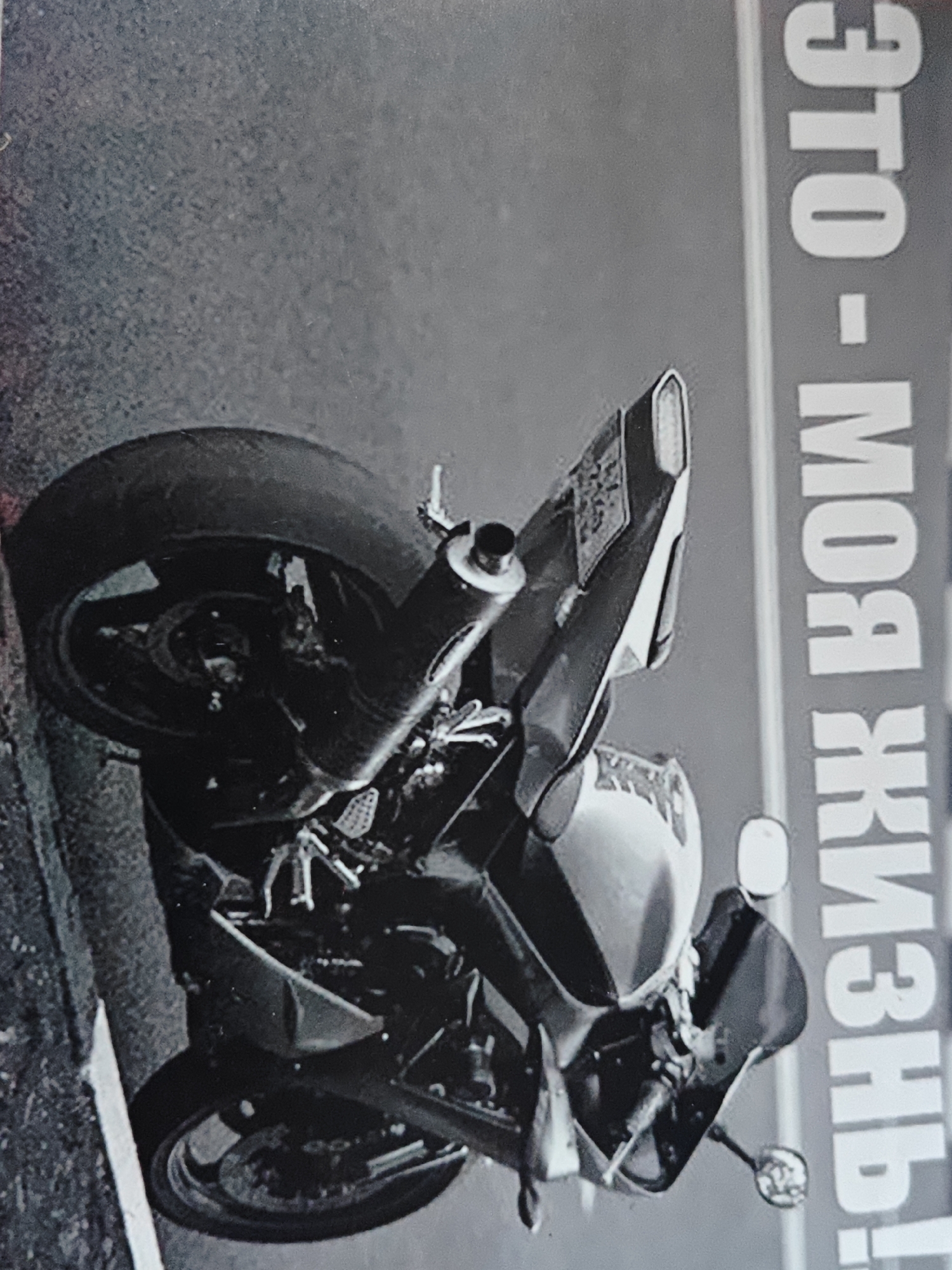 A black and white, low-angle, vertical shot captures a portion of a motorcycle parked on a textured, dark surface, possibly asphalt, in what appears to be an urban environment. The motorcycle is viewed from the rear, with its rear tire and exhaust system prominently featured. The tire is dark and appears worn. The exhaust pipe is visible on the right side of the motorcycle. To the right of the motorcycle, vertical white text is partially visible against a light gray background. The text appears to be in a foreign language and is positioned along the edge of the frame. The overall lighting suggests it is daytime. There are no people visible in the image.