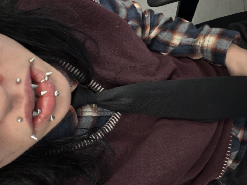 The image captures a close-up, downward-angled view of an individual's upper torso and lower facial region. The individual has long, dark hair. Their lips are slightly parted, displaying numerous metallic piercings and pointed, metallic, fang-like dental caps or veneers on the upper and lower canines. Specifically, there are two spherical piercings above the upper lip, four spiked piercings along the vermilion border of the upper lip, one central spherical piercing and one lateral spherical piercing on the lower lip, four spiked piercings along the vermilion border of the lower lip, and two additional spherical piercings below the vermilion border of the lower lip.
The individual is dressed in a multi-colored plaid shirt, featuring a pattern of dark blue, white, and reddish-orange squares, worn beneath a dark maroon or burgundy V-neck sweater or vest. The sweater/vest has a distinct white, horizontally striped trim along its V-neckline. A plain black, textured, narrow necktie is knotted at the collar and extends down the center of the chest. A dark-colored garment is partially visible at the bottom left of the frame. The background is an indistinct, light-toned indoor environment. A dark object, consistent with a chair or furniture component, is visible along the right edge of the image.
The individual is positioned as if looking directly into a camera. The image does not contain any discernible features or landmarks that would allow for the identification of a specific location within Sofia, Bulgaria.