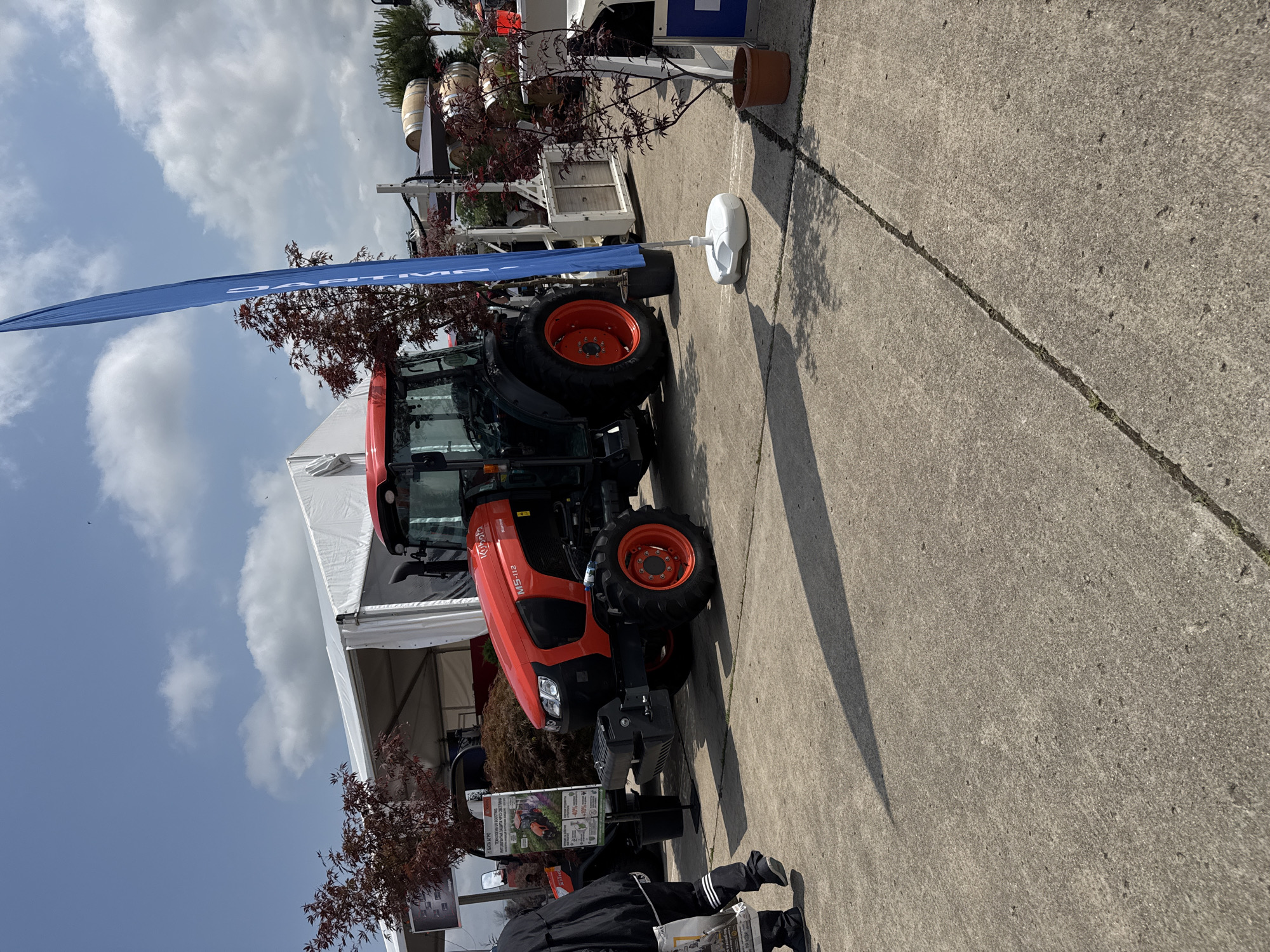 The image, taken outdoors in Stara Zagora, Bulgaria, displays a bright orange and black Kubota M5-112 tractor as the central subject. The tractor is positioned on a grey concrete surface, featuring large black tires with distinctive orange rims and a front-end loader attachment.
In the midground, to the left of the tractor, a tall blue flag or banner stands upright. Partially visible Cyrillic text on the flag suggests "PARTN". Behind the tractor and the flag, a large white tent is erected, likely part of an exhibition or display area. Scattered around the display are several potted plants with reddish-brown foliage. Further in the background, stacked wooden barrels and white industrial equipment are visible. The scene is set under a clear blue sky with scattered white clouds, indicating daylight hours.
In the bottom left corner, the lower body of a person is visible, wearing dark pants with white stripes and holding what appears to be a brochure or newspaper. An information stand with text and images related to agriculture is positioned near the tractor, partially obscured by the person. The concrete ground features a visible expansion joint. The overall setting suggests an outdoor agricultural fair or trade show, showcasing farm machinery and related products.