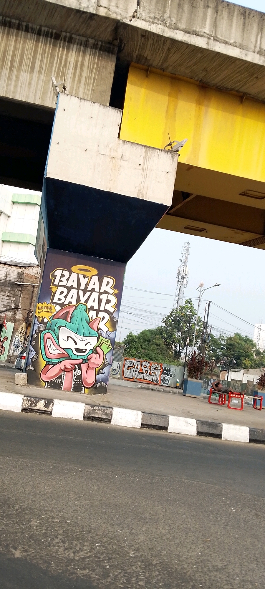 The image shows a street scene in Bekasi, Indonesia, under an elevated structure, likely an overpass or flyover. The pillar of the elevated structure is covered in graffiti. On the pillar, there is a cartoon character, possibly a cat or fox, depicted with a green hat and pink body, holding a banknote. Above the character, text in yellow with stars reads "BAYAR BAYAR 12". To the right of the pillar, a fence with more graffiti is visible, including a piece that appears to spell "FABS" in orange and white. A few trees and buildings are in the background, along with a telecommunications tower and a street lamp. In the middle ground, there is a sidewalk with benches, and a person is seen lying on one of the red benches. The foreground is dominated by an asphalt road with a white and black striped curb. The sky is overcast.