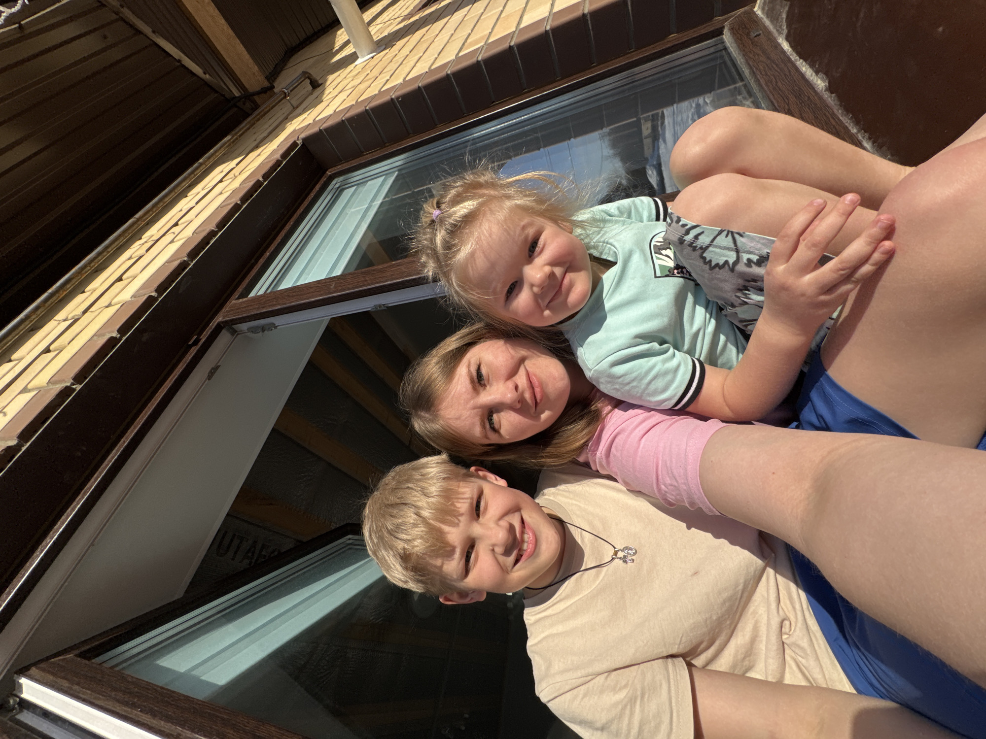 A photograph taken in Gornyi (Dvurechenskoe S/P), Russia, depicts three individuals positioned outdoors in direct sunlight. From left to right, an adolescent male, an adult female, and a young female child are visible, all smiling and facing the camera. The adolescent male has blond hair and light skin, wearing a light beige short-sleeved t-shirt and blue shorts. He wears a dark cord necklace with two small metallic pendants. The adult female, with light brown hair and light skin, is centrally positioned, wearing a pink long-sleeved top; she holds the young female child on her lap. The child, with blond hair gathered by a purple tie and light skin, wears a light blue short-sleeved t-shirt with a graphic print and patterned shorts. The subjects are situated in front of a building facade featuring dark brown window frames and large glass panes. These panes reflect a clear blue sky and a light-colored ground or distant landscape. Above the windows, light brown brickwork is visible, topped by dark brown architectural elements including an eave and an overhang. A white pipe is affixed to the brick wall on the right side of the frame. Strong shadows indicate bright, direct illumination.