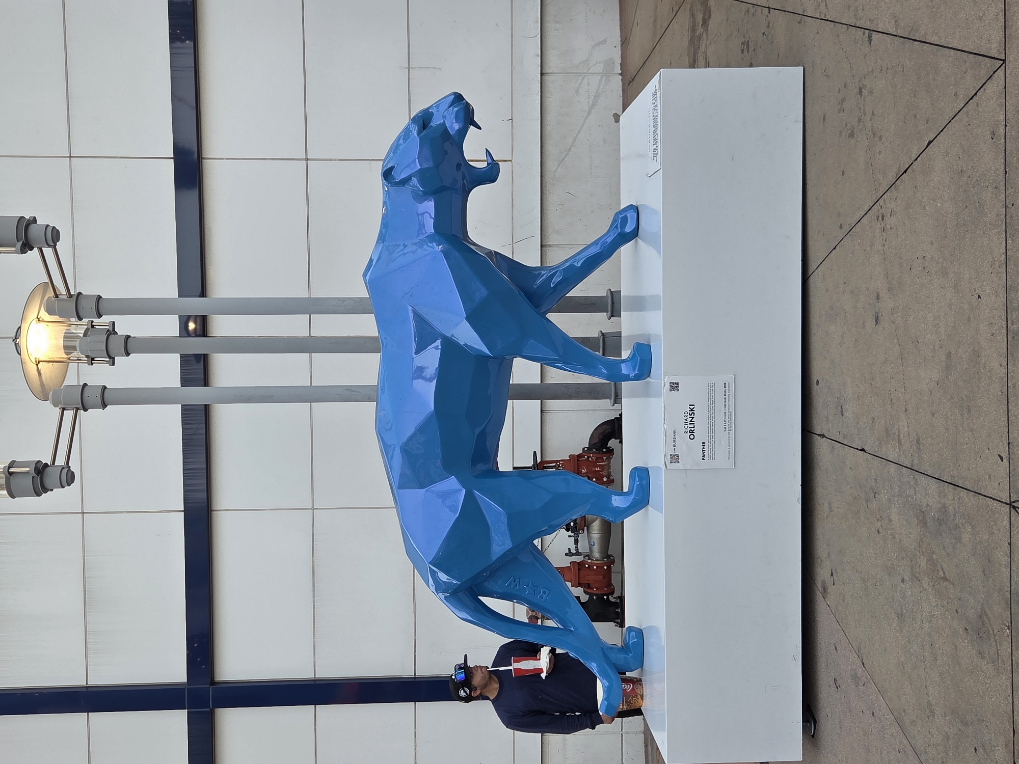 A blue, polygonal sculpture of a panther is displayed on a white pedestal in front of a white tiled building with a blue stripe. A man wearing a dark blue long-sleeved shirt, a baseball cap, sunglasses, and dark pants stands to the left of the sculpture. He is holding a red and white cup and a white plate with food. The sculpture is positioned in front of some red industrial pipes and machinery. A plaque is attached to the front of the pedestal, which reads "RICHARD ORLINSKI PANTHER". The background appears to be an urban environment, likely in Burbank, California, given the plaque's reference to Burbank. The scene is outdoors, with a concrete ground in the foreground. A lamppost with multiple lights is visible behind the sculpture.