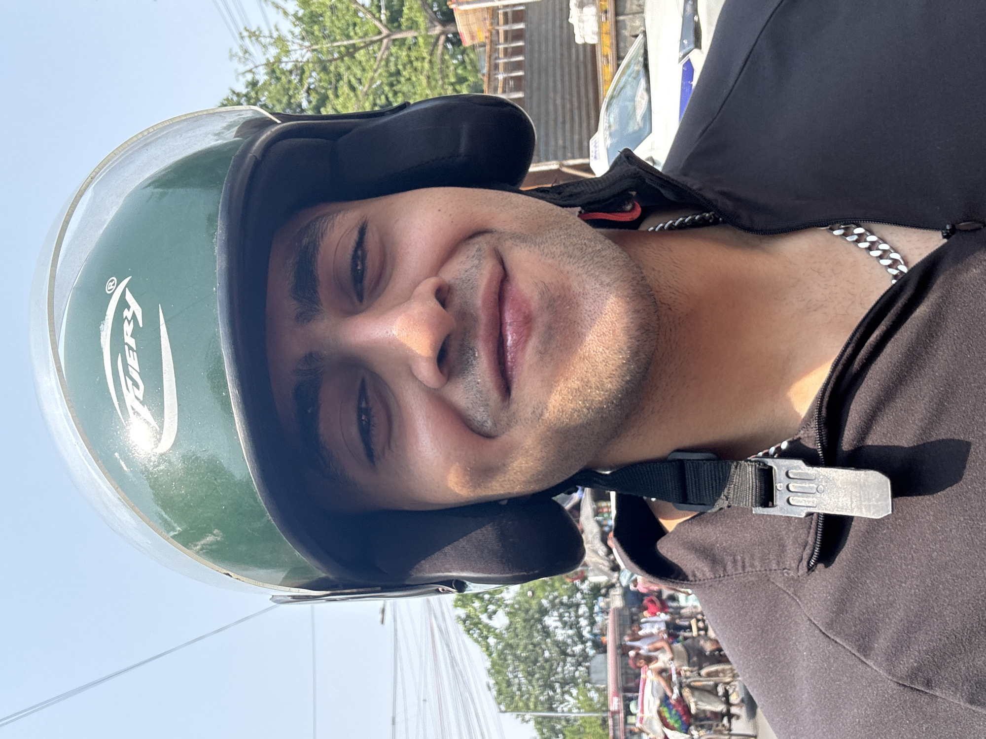 A man wearing a green helmet with a black liner and a black jacket poses for a photo. He is also wearing a silver chain. The helmet is fastened with a black strap and silver buckle. The background shows a crowded street scene, suggesting the photo was taken in Kolkata, India. Trees and power lines are visible against a bright blue sky.