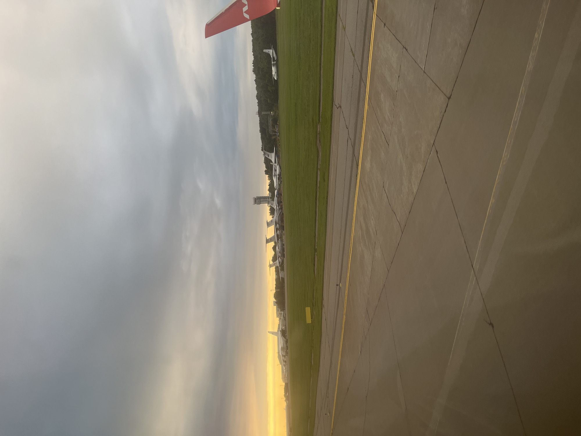 An aerial view captures an airport tarmac and grassy field under a cloudy sky with the sun setting on the left, casting a warm glow. Multiple airplanes are parked on the grass, with a control tower visible in the distance. A portion of an airplane wing, painted red with a white logo, is in the foreground on the right. The scene suggests a twilight departure or arrival at an airport, possibly in Stolbishche, Russia, given the context.