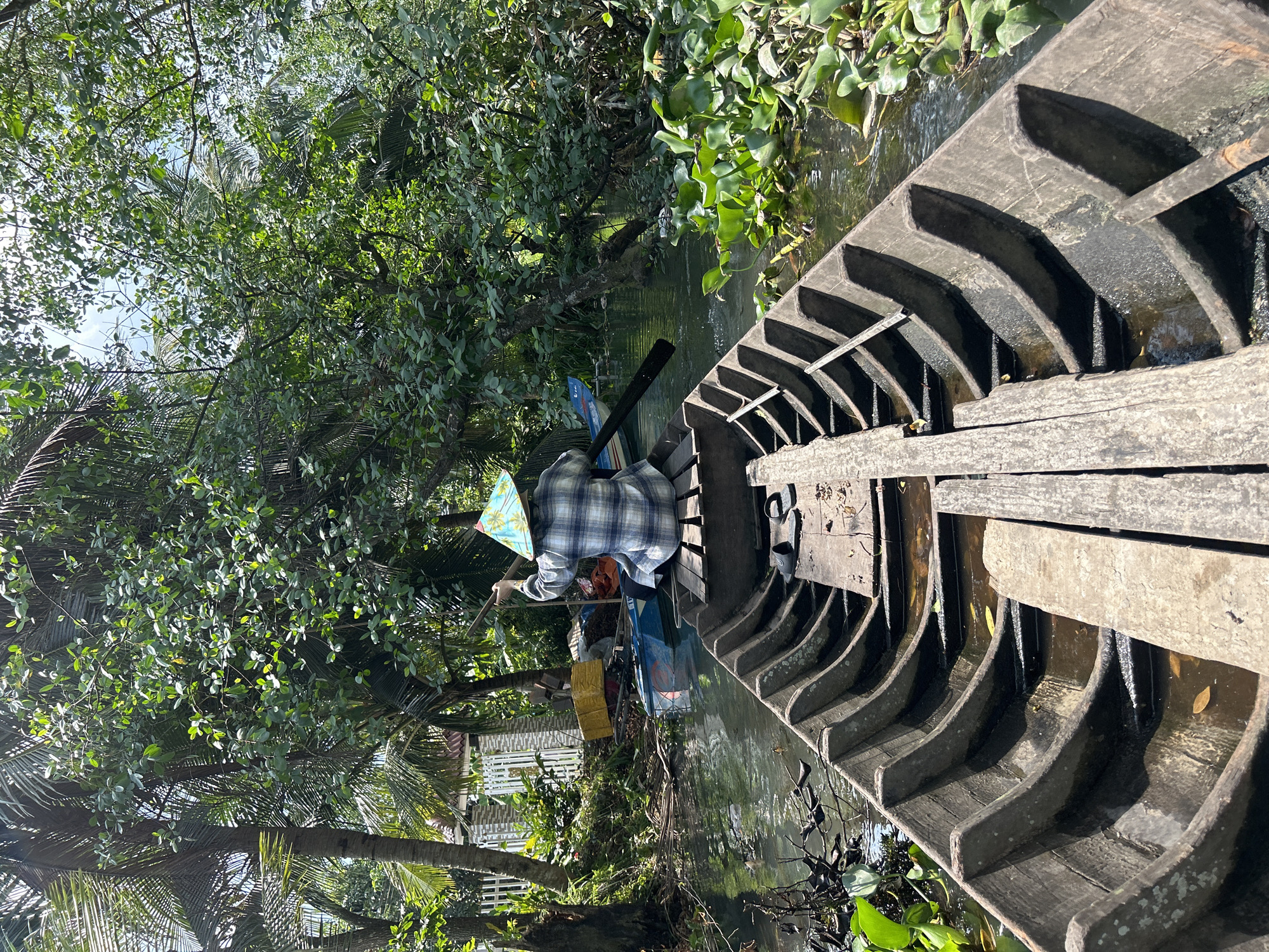 A person wearing a blue and white plaid shirt and a conical hat is rowing a wooden boat down a narrow canal. The canal is lined with lush green trees and vegetation. In the background, there is a house with a white fence and a blue boat. The water is dark and reflects the surrounding greenery. The scene appears to be in a rural area of An Hữu, Vietnam, likely a canal or waterway common in the region's rural landscape. The image captures a serene moment of traditional transportation and life along the waterways.