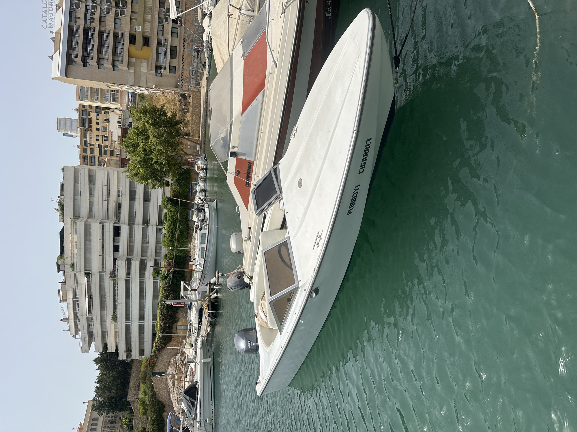 In Palma, Spain, a white speedboat with the registration PL0003YI and the name "CIGARRET" printed on its side is docked in a harbor. The boat features two outboard motors, one of which is clearly visible with the number "40" on its casing. The boat has a tan-colored interior with a windshield. Adjacent to this boat, another larger boat with a red and white striped cover is moored. In the background, several buildings, including a multi-story white apartment building with balconies and a taller beige building with balconies, are visible. The water is a clear, turquoise green, and a rope is tied to the front of the white speedboat.
