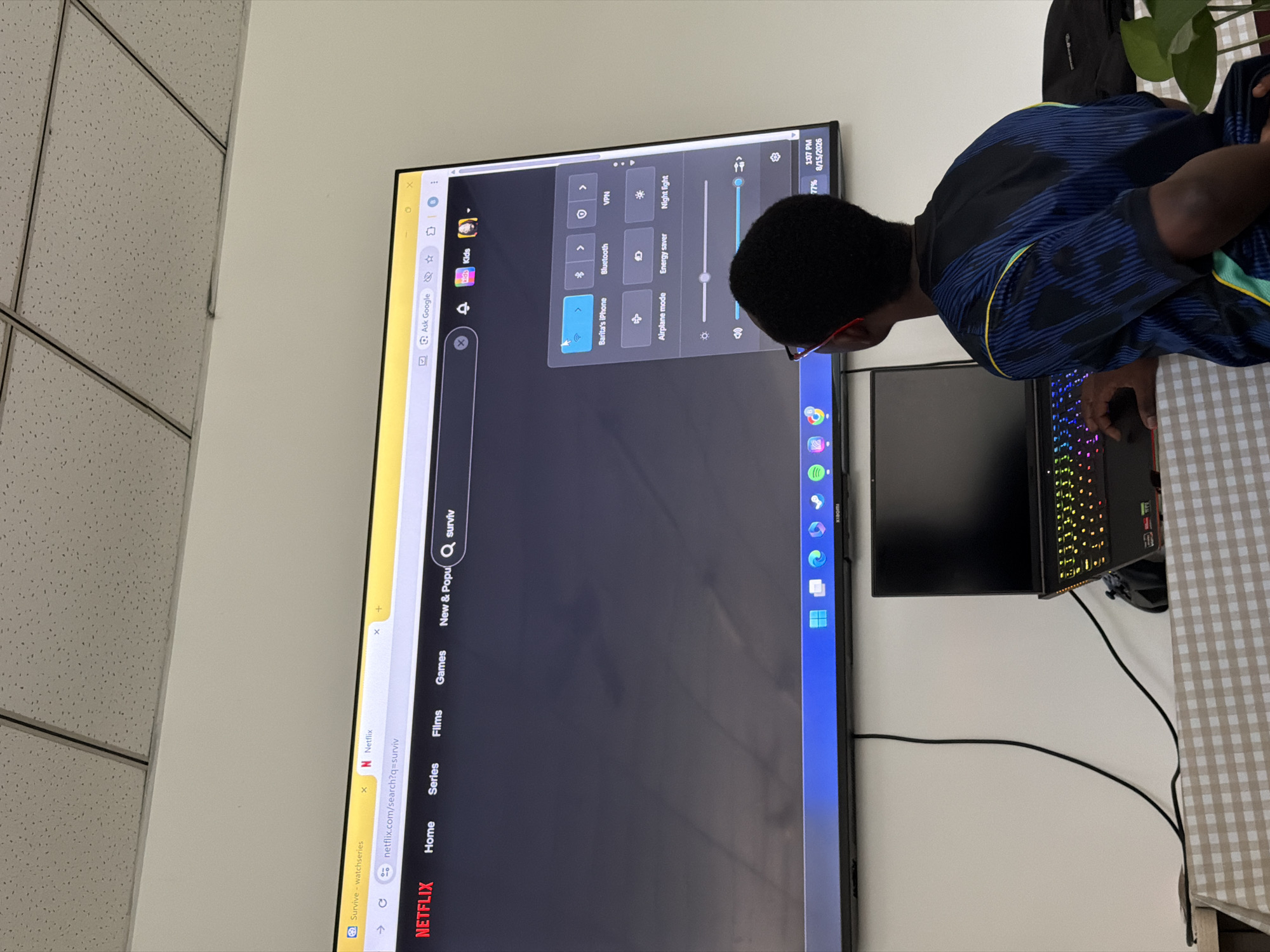 The image shows a young Black male with glasses wearing a blue and yellow shirt, sitting in front of a large screen television. The television displays a Netflix search results page for "surviv" and a settings menu with options like "Barita's iPhone," "Bluetooth," "VPN," "Airplane mode," "Energy saver," and "Night light." There are also sliders for brightness and volume. A laptop with a colorful, illuminated keyboard is placed on a table in front of the television. The room appears to be an indoor space with a drop ceiling. The overall impression is of a person using technology, likely for entertainment or work. There is no indication of the location being in Jiangjin, China.