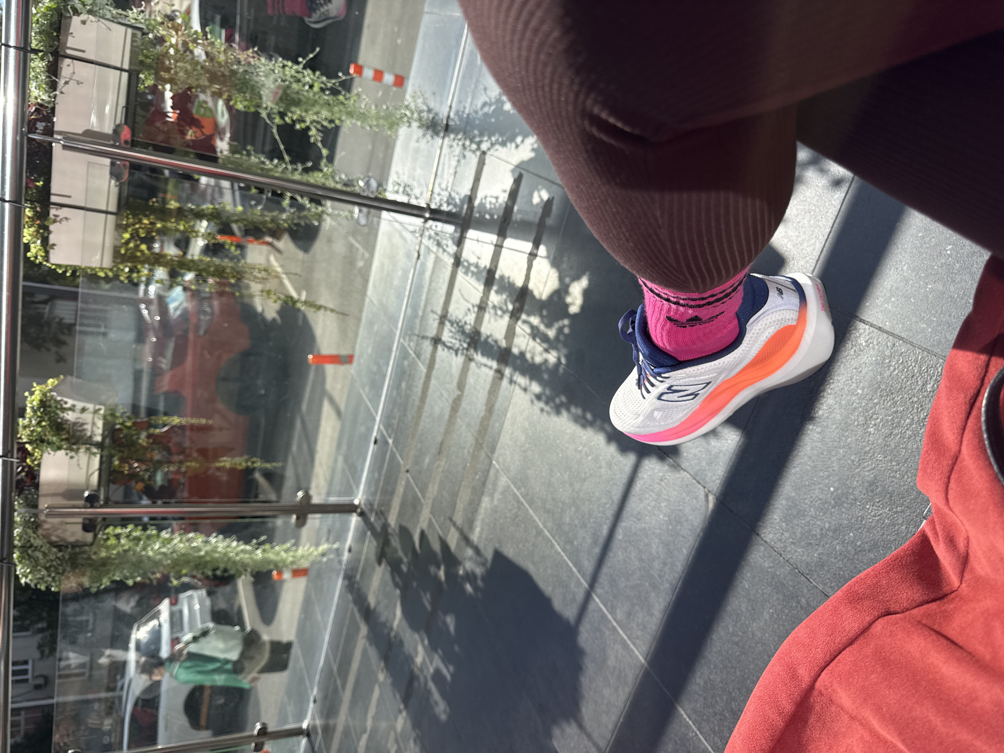 The image captures a low-angle, close-up perspective of a person's right leg, adorned with a vibrant pink sock and a white athletic shoe with orange and blue accents, resting on a dark tiled floor. The shoe features the New Balance logo. The person is wearing dark, ribbed leggings. A portion of a red garment, possibly a jacket or bag, is visible in the lower left corner.
The background suggests an outdoor urban setting, likely a cafe or public space with a glass barrier. Through the glass, cars and buildings are visible. A white SUV and a red car are prominent. In the distance, a person in a green jacket is walking. Orange traffic cones are placed along the walkway outside. Hanging plants, some trailing green vines, are suspended from planters above the glass barrier. The sunlight casts long shadows of the plants and the glass barrier onto the tiled floor, indicating a sunny day. The architecture visible in the background, particularly the building on the left with multiple windows and balconies, is consistent with residential or commercial buildings found in cities like Moscow, Russia. The overall impression is of a casual outdoor scene in a city environment.