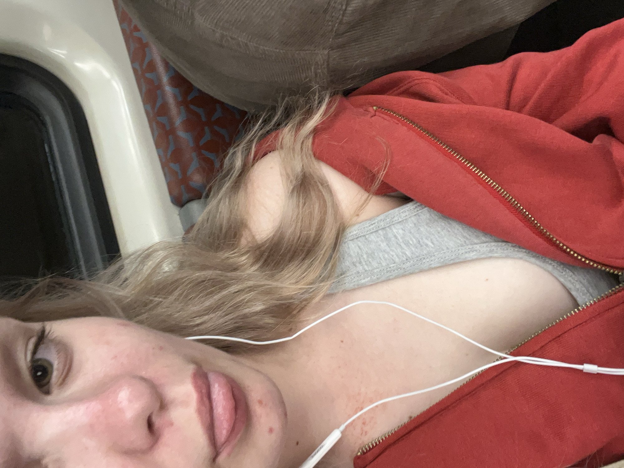 The image depicts a close-up view of a person, likely female, sitting on public transportation. The subject has fair skin, blonde hair, and is wearing a red zip-up hoodie over a grey tank top. White earphones are visible. The background shows the interior of a vehicle, possibly a train or bus, with a window on the left and patterned seating in red and blue. A portion of another passenger's brown corduroy clothing is visible on the right. The lighting suggests an indoor, potentially dim environment. The presence of a window and seating pattern implies a public transit setting. There is no information to place this scene specifically in Saint Petersburg, Russia.