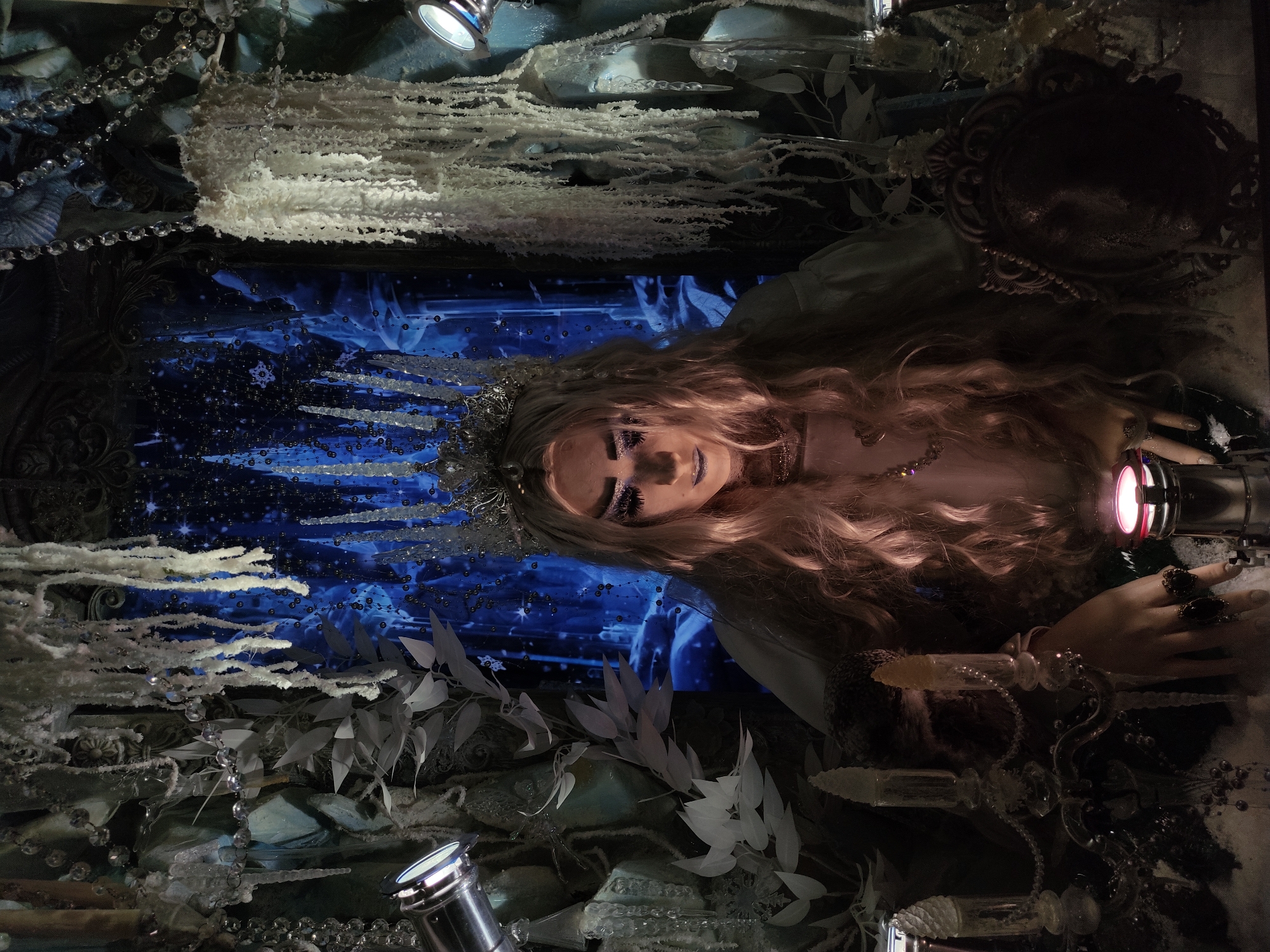 A dimly lit, ornately arranged window display features a mannequin with blonde hair, wearing a crown and elaborate makeup. The mannequin is positioned horizontally and is adorned with jewelry. The backdrop is a blue and black pattern with apparent beads and crystals hanging. There are also decorations including branches, crystals, and light fixtures visible in the scene. The location of the display is unknown.