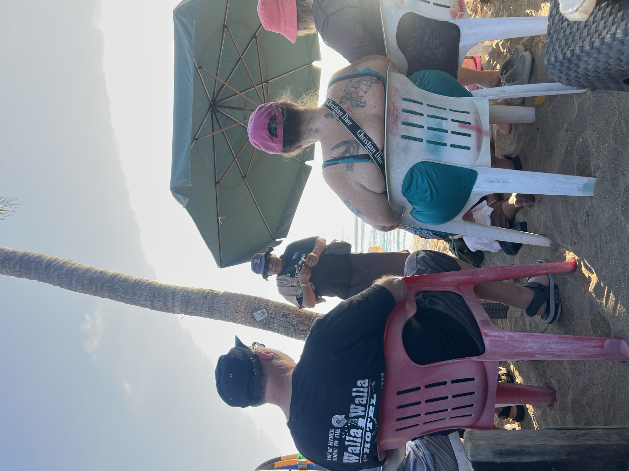 In Kuta, Indonesia, on a sandy beach, a group of people are seated, facing away from the camera. In the foreground, a man wearing a black t-shirt with "Walla Walla Hotel" printed on the back sits in a red plastic chair. Next to him, to his right, a woman with a pink hat and a teal swimsuit sits in a white plastic chair. She has multiple tattoos on her back and a Christian Dior strap across her chest. To her right, another person wearing a pink hat sits further back, partially obscured. In the middle ground, a man wearing a blue bucket hat and sunglasses stands, holding a camera or similar device. He appears to be interacting with the seated individuals. A large, green umbrella is open, providing shade. A palm tree trunk is visible to the left of the umbrella. The sun is setting, casting a warm glow and creating lens flare. The environment is a beach, indicated by the sand and the ocean in the background.