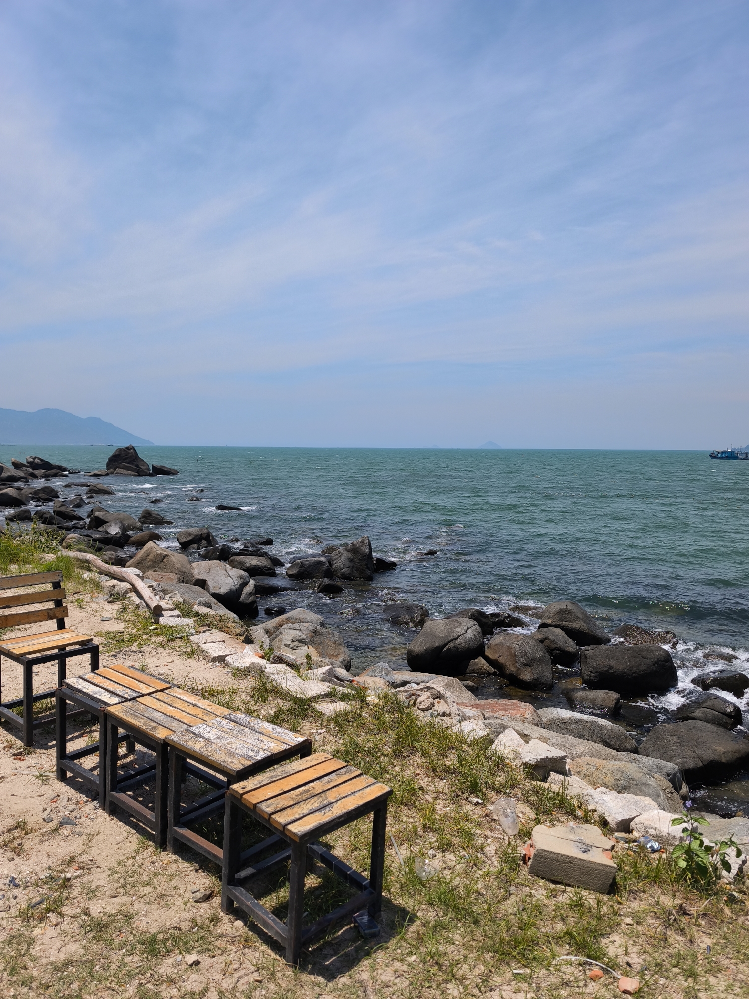 An outdoor scenic view captured on a clear day shows a rocky coastline with benches in the foreground. The sea stretches out to the horizon, with a faint mountain range visible in the distance on the left and a boat on the right. The foreground consists of sandy ground with sparse vegetation, leading to a rocky shoreline. Two wooden benches with metal frames are positioned on the sand. The benches are rustic in appearance, with weathered wooden slats. The rocky shore is covered in various sizes of grey and brown stones, with some rocks submerged in the water. The sea is a calm, deep teal color, with small waves lapping against the shore. The sky is a pale blue with wispy white clouds. The overall scene suggests a tranquil coastal setting, potentially a public park or a quiet spot for observation.