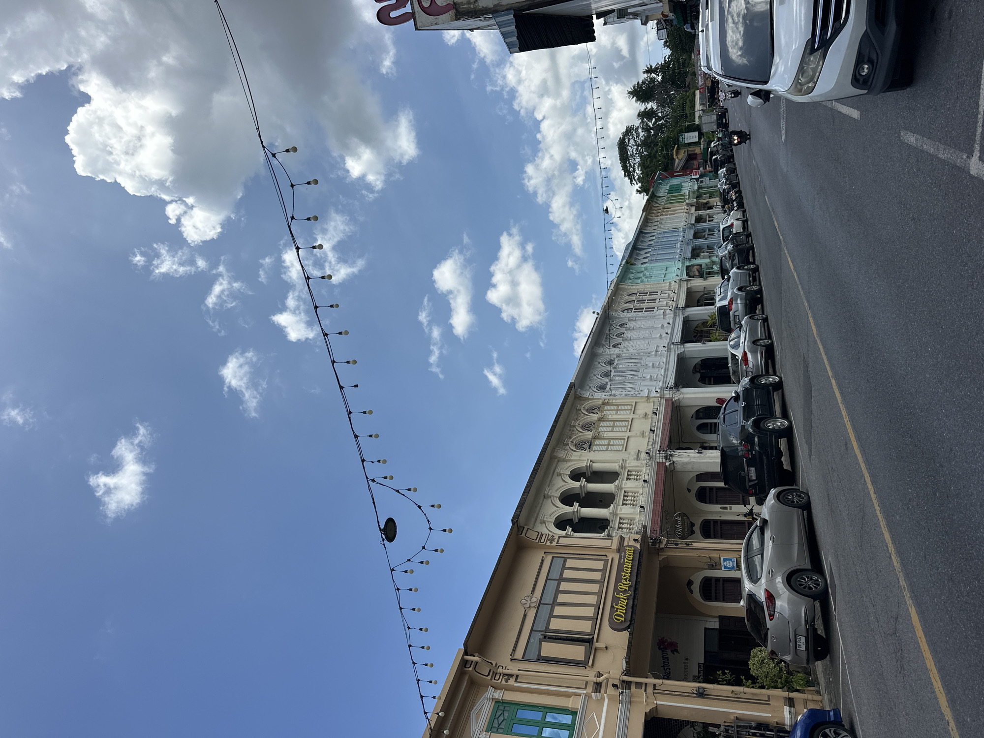 The image depicts a street scene in Phuket City Municipality, Thailand, characterized by a row of colorful heritage buildings lining the right side of the road. The architecture features distinct colonial influences with ornate balconies, arched windows, and pastel-colored facades. String lights with exposed bulbs are suspended across the street, illuminating the scene. Parked cars, including a silver hatchback, a black SUV, and a white SUV in the foreground, occupy the parking spaces along the buildings. The road itself is asphalt, marked with a yellow line. In the distance, lush green trees and a hillside are visible. The sky is a bright blue with scattered white cumulus clouds. A few motorcycles are also present on the road. The overall impression is that of a vibrant, historic street with elements of both traditional and modern life.