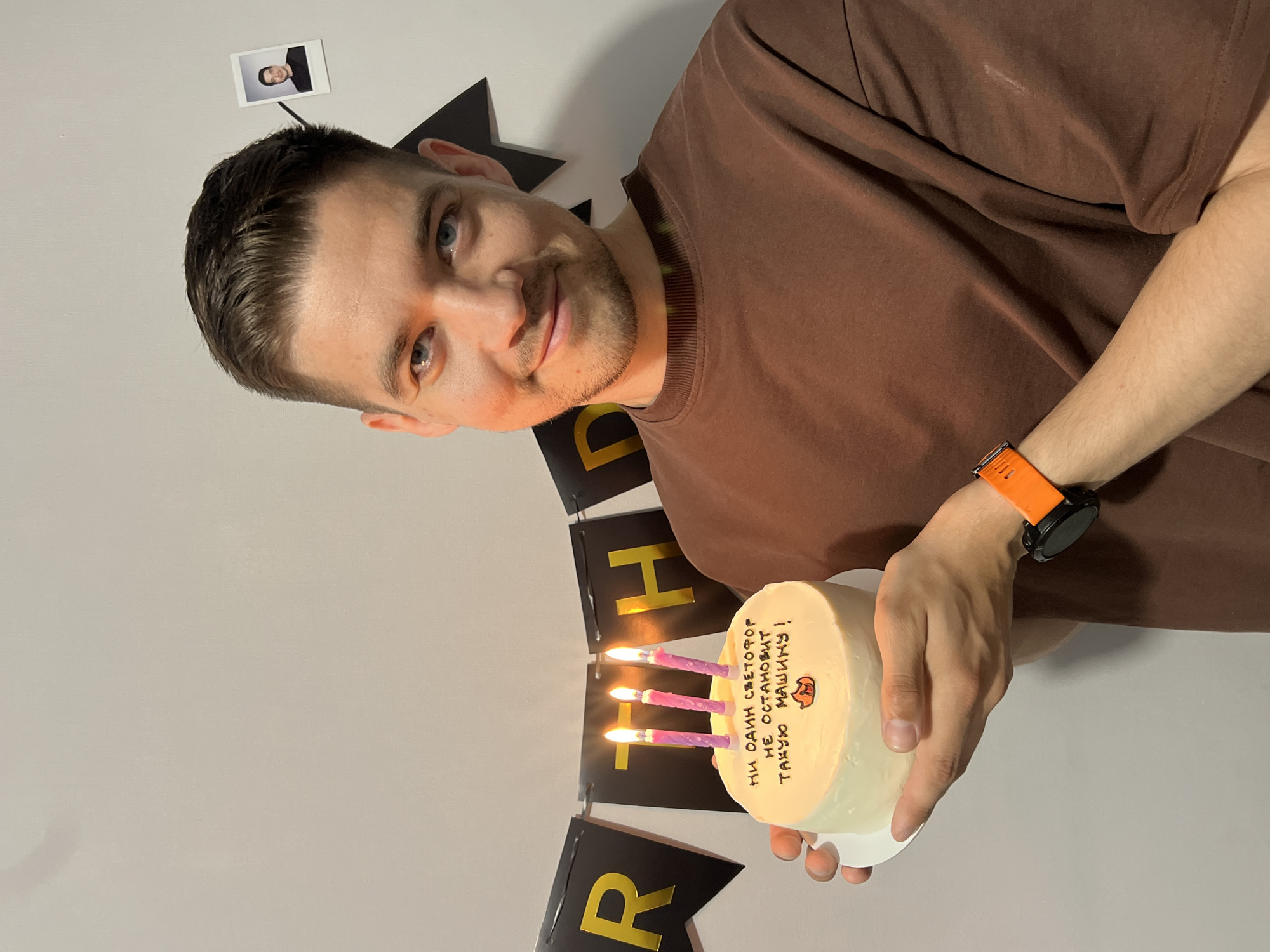 A man is holding a birthday cake with candles in front of a "BIRTH" banner. The cake has writing in Russian and a small drawing of an animal on it. The man is wearing a brown shirt and an orange smartwatch. There is a photo attached to the wall behind him. The background is a plain, light-colored wall. There is no indication of Kazan, Russia, from this image.
