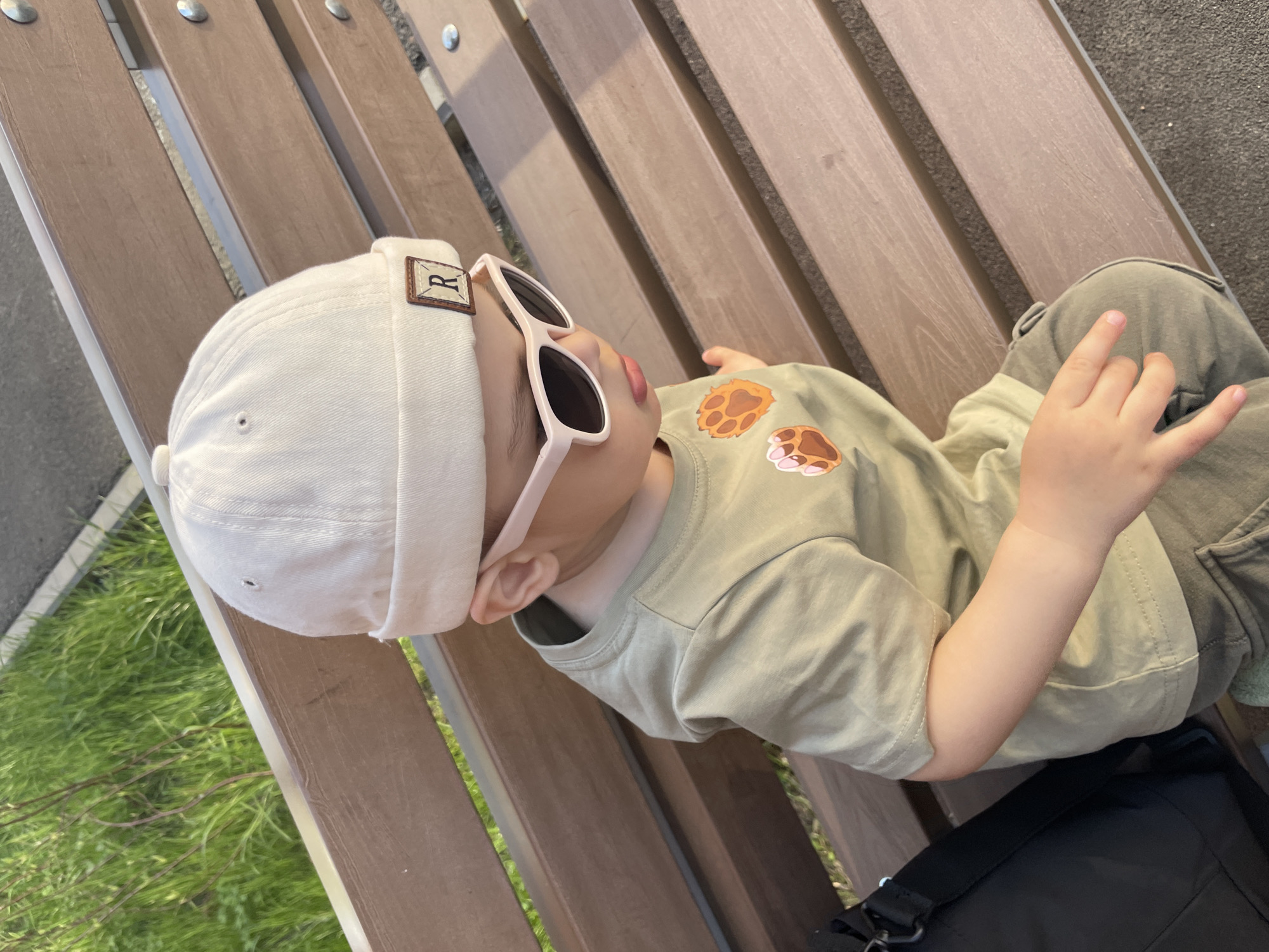 A child is lying on a wooden bench outdoors. The child is wearing a white cap with a patch, pink sunglasses, and an olive green t-shirt with paw print designs. The child is also wearing green cargo pants. The child's hand is raised slightly. A black bag rests on the bench near the child. Green grass is visible in the background. The location is undetermined.