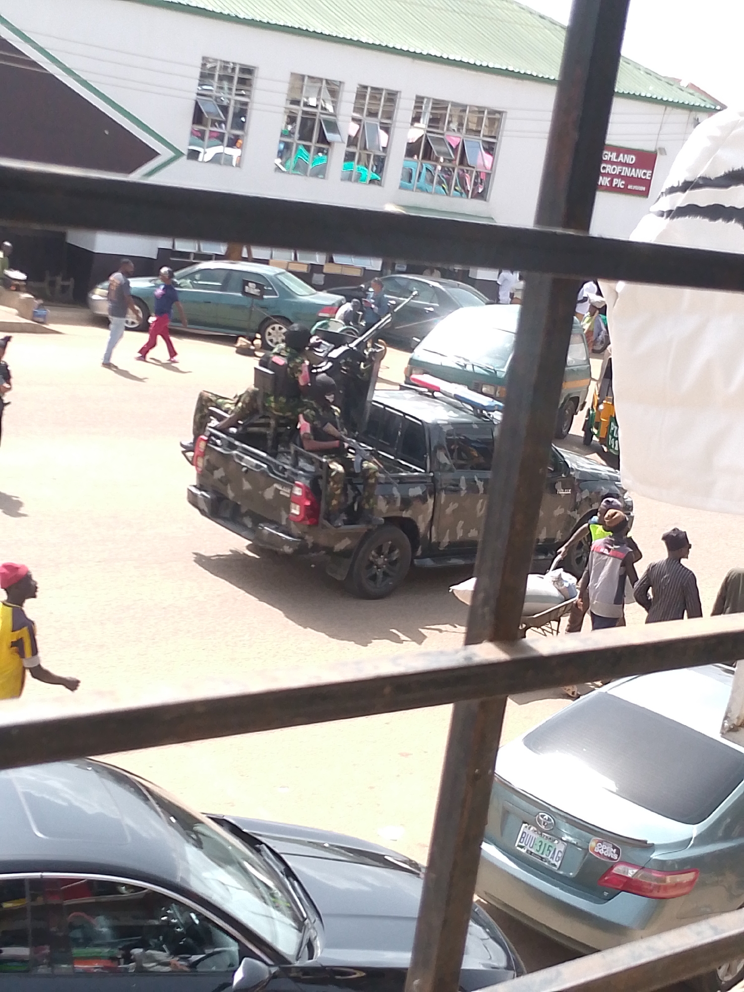 In Bukuru, Nigeria, a light-colored Toyota Camry, license plate BUU-315AG, is parked in front of a building with a green roof and white facade. The building has several windows with tinted glass reflecting the sky. A sign on the building reads "HIGHLAND CROFINANCE NK Plc." In the foreground, a dark-colored truck with camouflage paint is parked, containing several individuals in military-style uniforms armed with rifles. To the left of the truck, a man in a yellow and red shirt and a red cap is walking. Other individuals are visible in the background, including two walking near a green sedan, and others near the truck and a three-wheeled vehicle. The scene appears to be a public street or parking area.