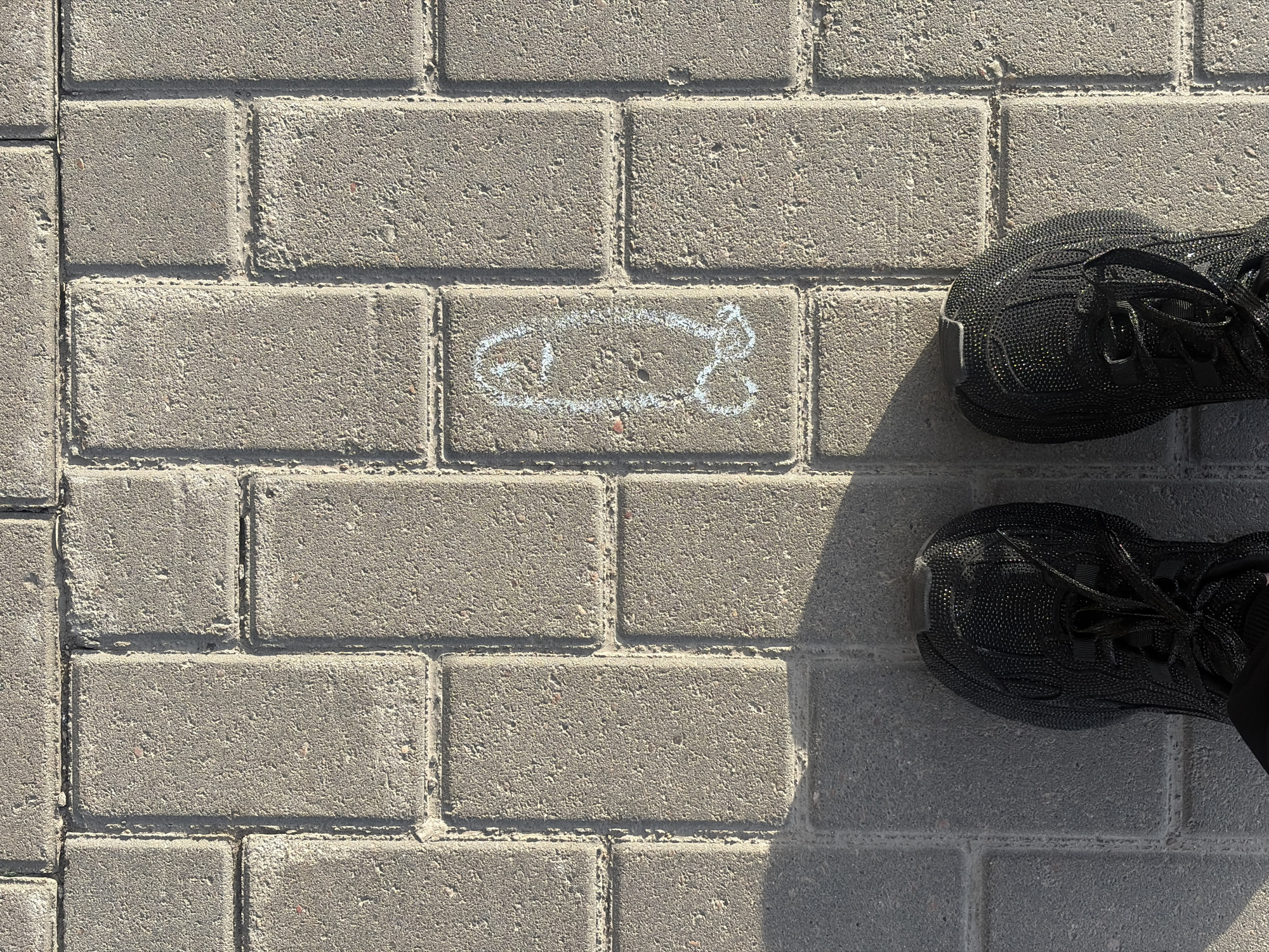 The image shows a section of paved walkway made of rectangular gray pavers. On one of the pavers, a simple outline of a fish is drawn in white chalk. A pair of black running shoes are partially visible in the upper right corner of the image. The location is the city of Saint Petersburg, Russia.