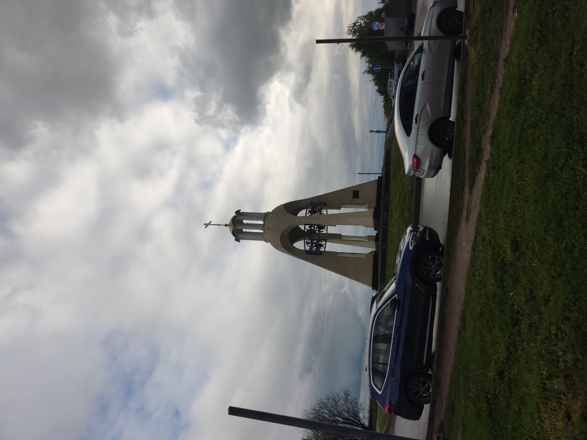 The image depicts a bell tower situated in the city of Koltushi, Russia. The tower is constructed with light-colored material and features a cross at its apex. There are bells visible within the tower's structure. The sky is overcast with a variety of cloud formations. On the right side of the image, along the roadside, two cars are parked. A blue SUV is closer to the viewer, while a silver sedan is positioned further away. Vegetation, mainly grass, is visible along the edge of the road. Street lighting and road signs are present near the vehicles.