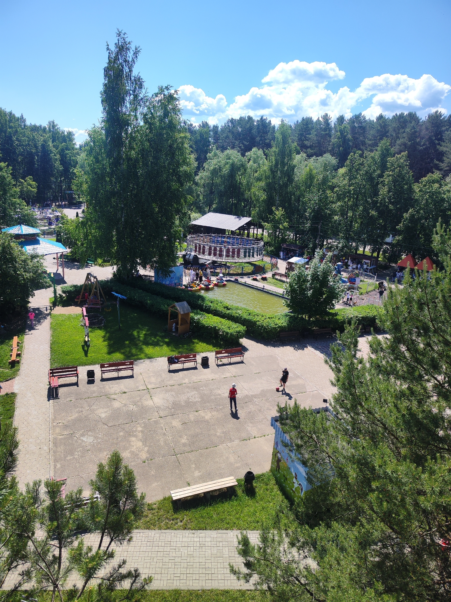 An aerial view depicts a park area on a sunny day with scattered clouds. In the foreground, pine trees obscure parts of the scene. A concrete pathway leads through the park, bordered by green grass. Several red benches and black trash cans are placed along the pathway. Further into the park, a grassy area with a playground set, including swings and a small wooden playhouse, is visible. Adjacent to this is a body of green water with several paddle boats, and a circular ride structure with a covered roof. A large building with a dark roof and a white awning is situated near the water. In the middle ground, two individuals are present on the concrete area; one person is wearing a red shirt and white hat, standing near a red ball, while another person in a black shirt and shorts is kicking the ball. A building with colorful murals on its side is also present. The background is dominated by a dense forest of deciduous and coniferous trees.