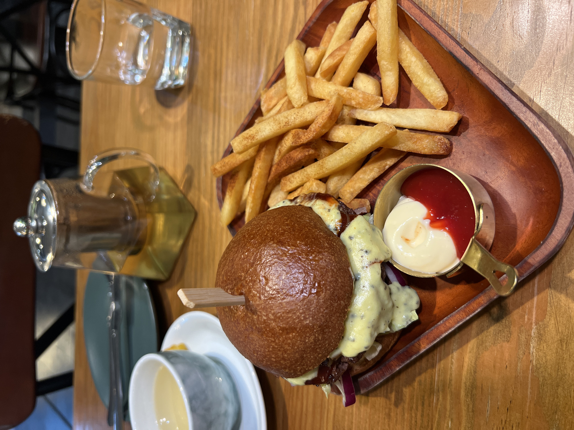 On a wooden table in what appears to be a restaurant, there is a wooden platter featuring a burger, fries, and a small metal container with two sauces (ketchup and mayonnaise). Next to the platter, there's a clear glass with what looks like a water pitcher with a brass base. Nearby, there's a small cup on a saucer.
It is not possible to discern the location being in Nha Trang, Vietnam from the visual information provided.