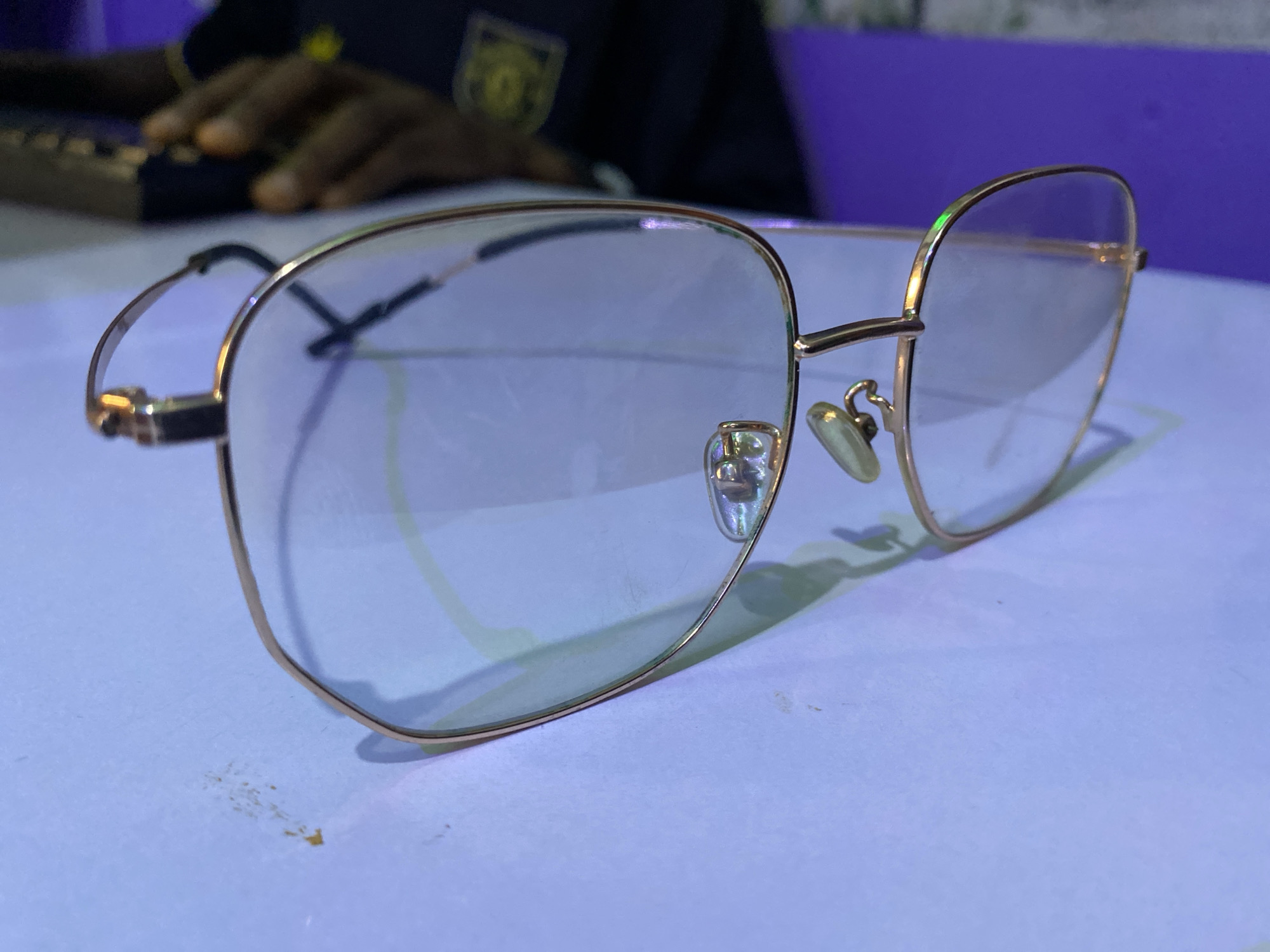 The image shows a pair of gold-framed aviator eyeglasses resting on a light blue or white surface. The glasses are slightly angled, casting a shadow on the surface. In the background, there is a blurred figure with dark skin using a computer keyboard, with what appears to be a Manchester United crest visible on the person's shirt. The environment is indoor, possibly an office or home setting. There is no information available to determine if the location is Bukuru, Nigeria.