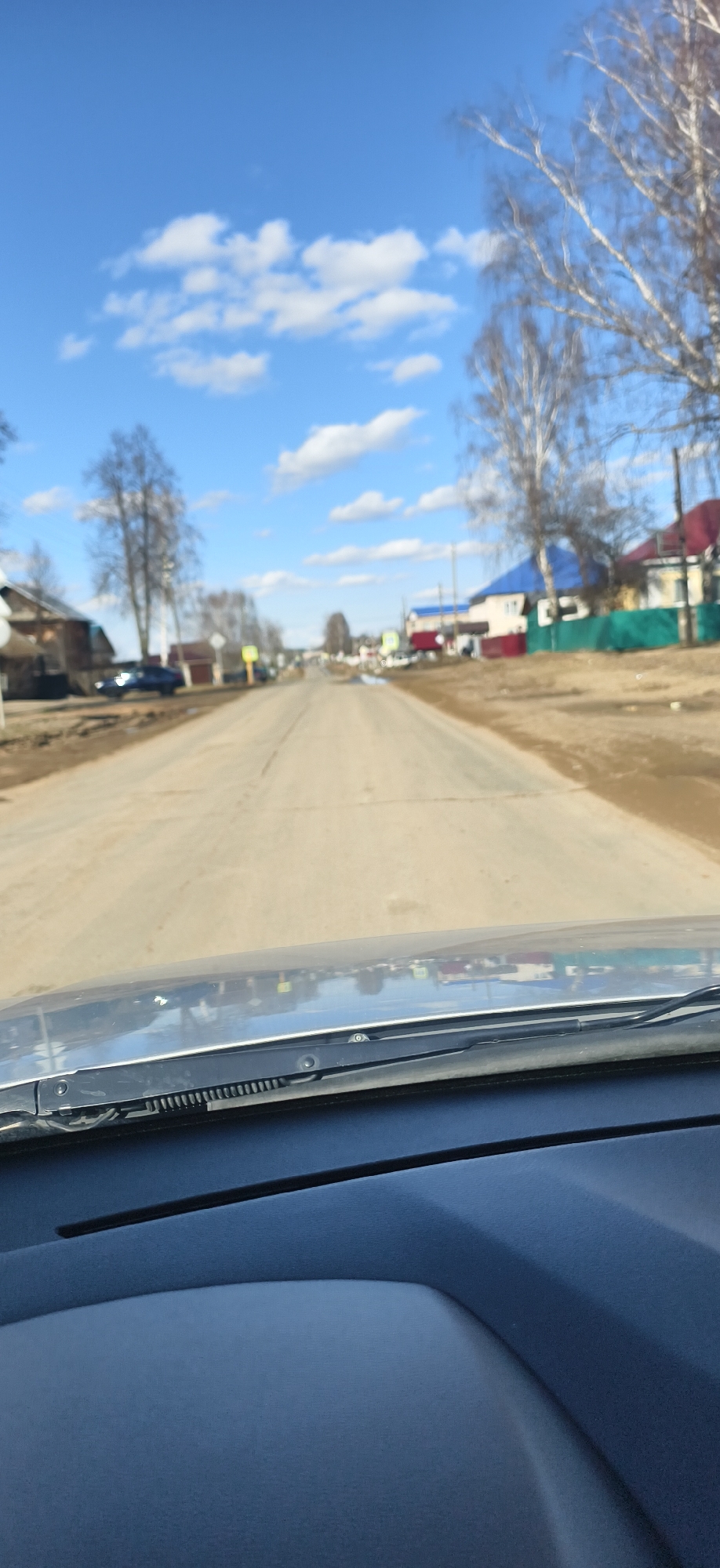 The image shows a view from inside a vehicle, likely a car, looking down a straight, paved road in Фоки, Russia. The road is flanked by houses and trees with bare branches, suggesting early spring or late fall. The sky is blue with scattered white clouds. The dashboard of the vehicle is visible in the foreground, including a windshield wiper.
