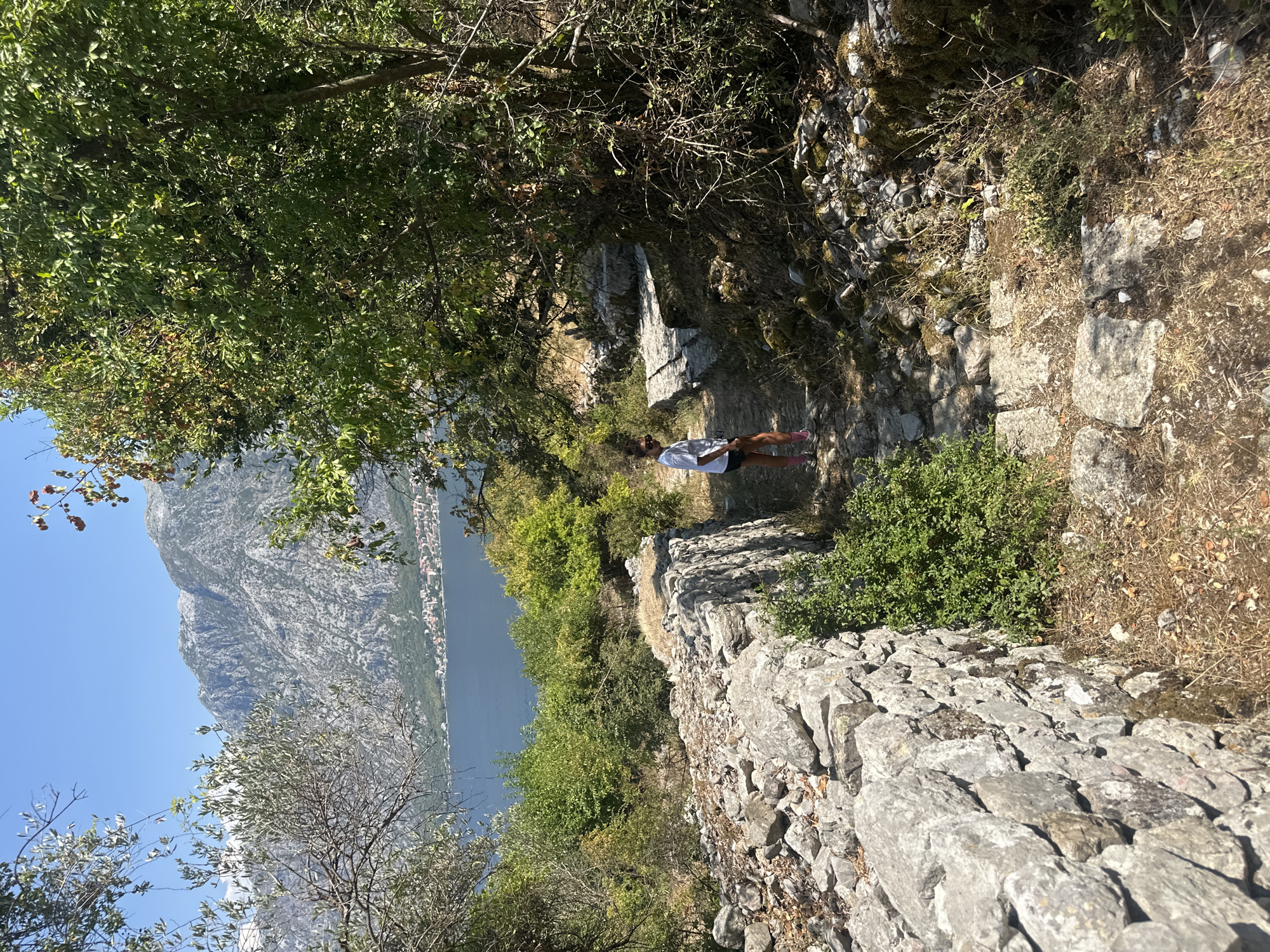 In Prčanj, Montenegro, a young person wearing a white t-shirt, dark shorts, sunglasses, and pink socks stands on a stone path. The path is bordered by a rough stone wall on the left and lush green trees on the right. In the background, a vast blue body of water, likely the Bay of Kotor, is visible, with a town nestled along its edge and majestic, rocky mountains rising behind it under a clear blue sky. The scene is bathed in bright sunlight, casting shadows on the path and highlighting the textures of the stone and foliage.