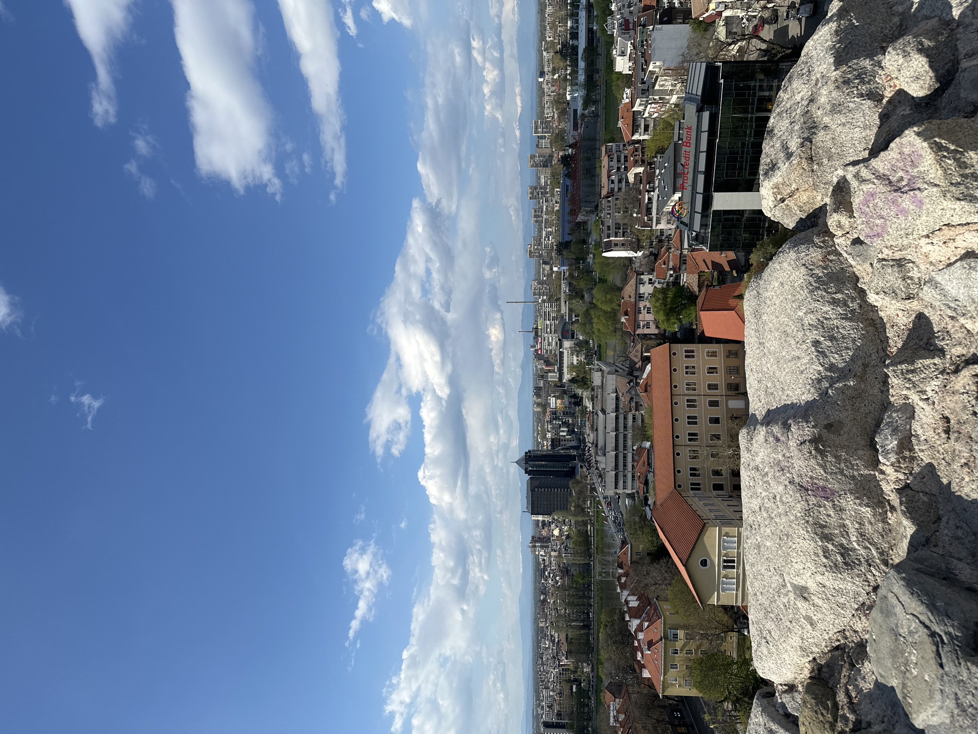 The image captures a view from atop a rocky outcrop overlooking the city of Plovdiv, Bulgaria. The sky occupies approximately half of the frame, filled with bright blue and elongated white clouds. Below, the city stretches towards the horizon, a mix of residential buildings, commercial structures, and greenery. A river winds through the urban landscape. The foreground is dominated by large, light-colored rocks, likely part of the elevated viewpoint.