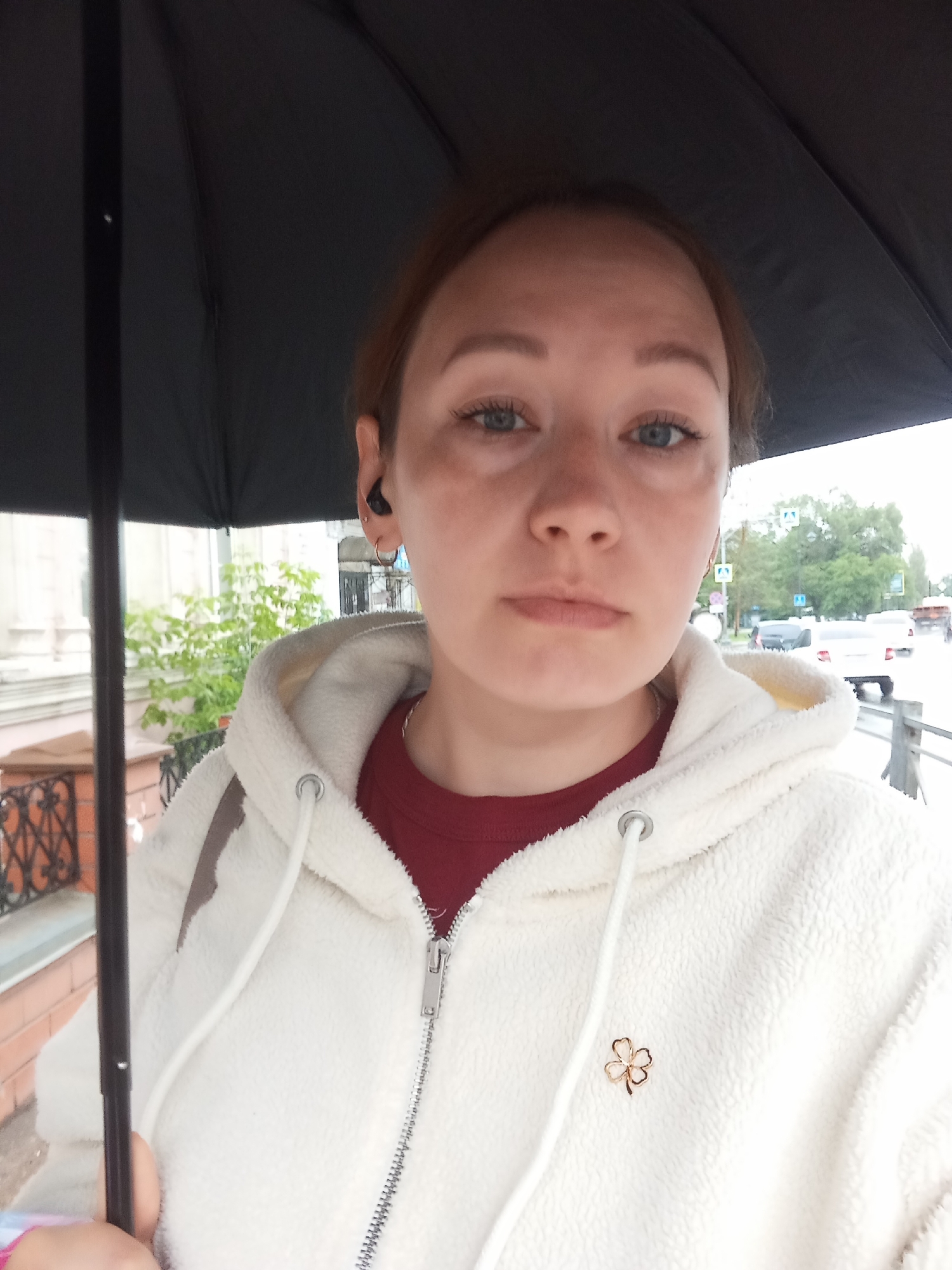 A medium-shot, eye-level selfie shows a young woman holding a black umbrella over her head, shielding her from the rain. She is wearing a cream-colored hooded fleece jacket over a maroon t-shirt. The woman has light skin, light brown hair, and blue eyes, and is looking directly at the camera with a neutral expression. A small gold clover-shaped pin is attached to the left side of her jacket. Her left ear has a small gold hoop earring and a black wireless earbud. In the background, across a street, a green tree and building facade are visible, with traffic signs and cars on a wet road, suggesting an urban environment in Orenburg, Russia. The overall lighting is subdued, consistent with overcast weather.