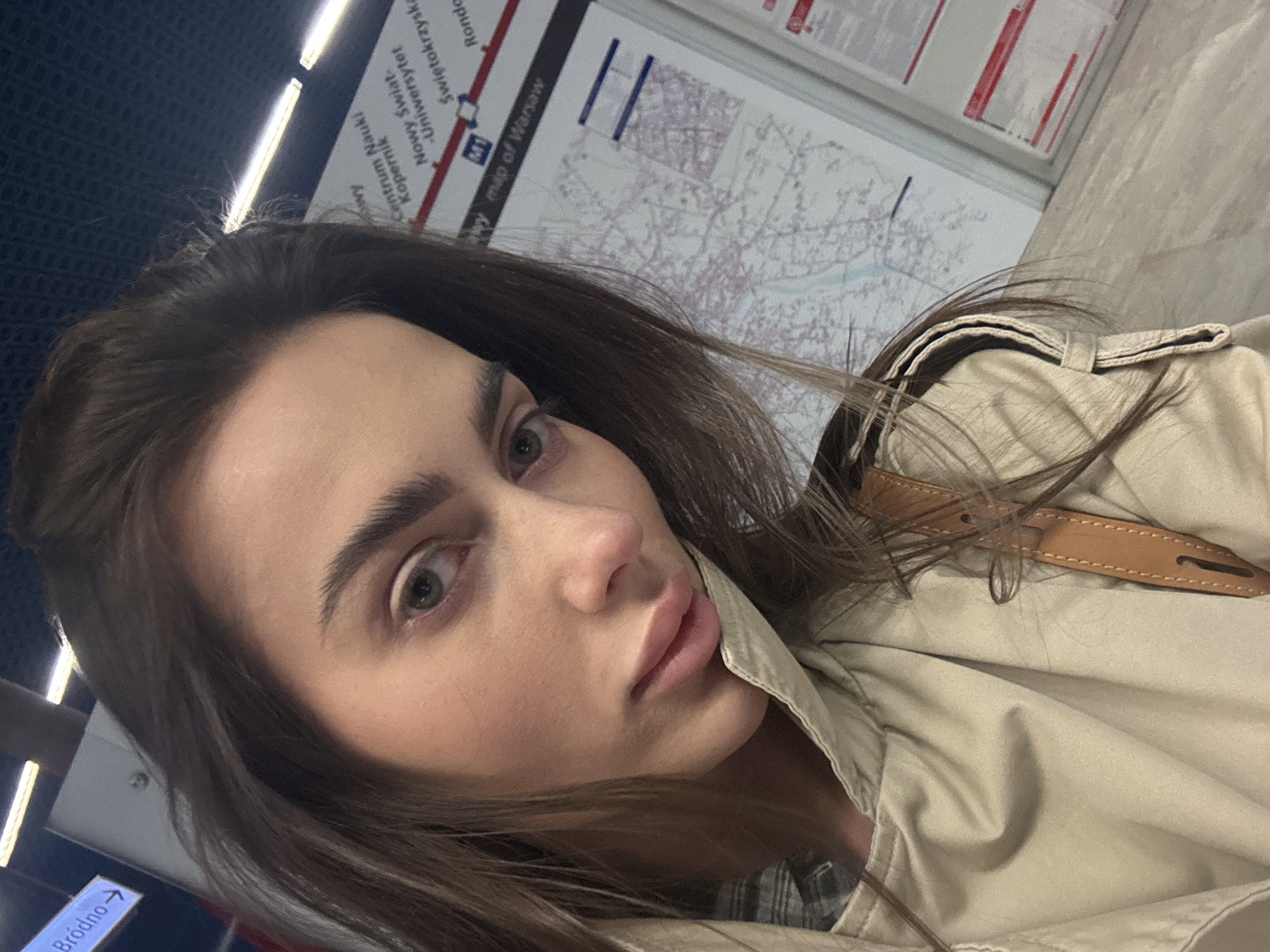 The image is a close-up, tilted selfie of a young woman taken inside what appears to be a subway station or train in Warsaw, Poland. The woman, who is the primary subject, is positioned off-center, facing slightly to her right. She has long, dark brown hair, prominent eyebrows, and light-colored eyes. Her facial expression is neutral and composed. She is wearing a light beige or tan trench coat, and a brown leather strap, possibly from a bag, is visible over her right shoulder.
The background provides strong contextual details for the location. Behind her, a large poster displaying a "map of Warsaw" is clearly visible, showing routes and various station names. Legible names on the map include "Świętokrzyska", "Rondo", "Centrum Nauki Kopernik", and "Nowy Świat-Uniwersytet", along with the "M1" metro line symbol, confirming the setting as the Warsaw Metro system. In the lower-left corner, an illuminated sign points to "Bródno→", indicating a direction or destination.
The environment features artificial lighting, with linear light fixtures visible in the upper part of the frame. The walls appear to be a mix of dark blue, textured paneling on the left and lighter, possibly concrete or tiled surfaces on the right. The overall impression is an everyday moment captured within a public transportation setting.